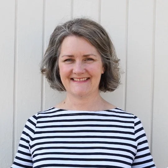 Woman with gray, wavy hair smiling in front of a white wooden wall, wearing a black and white striped shirt.