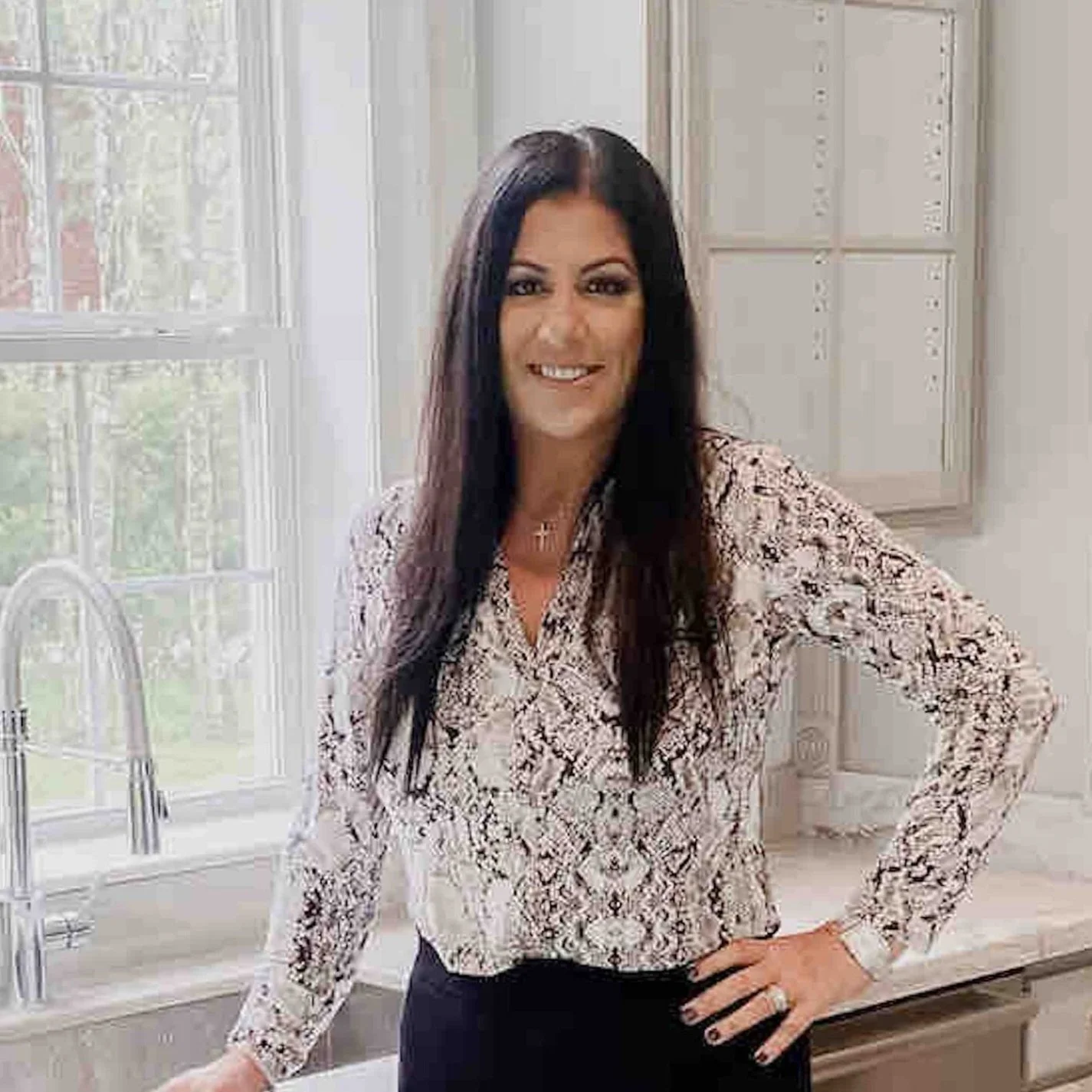 Woman with long dark hair wearing a snake print blouse, standing in a bright kitchen with a window, smiling with hand on hip.