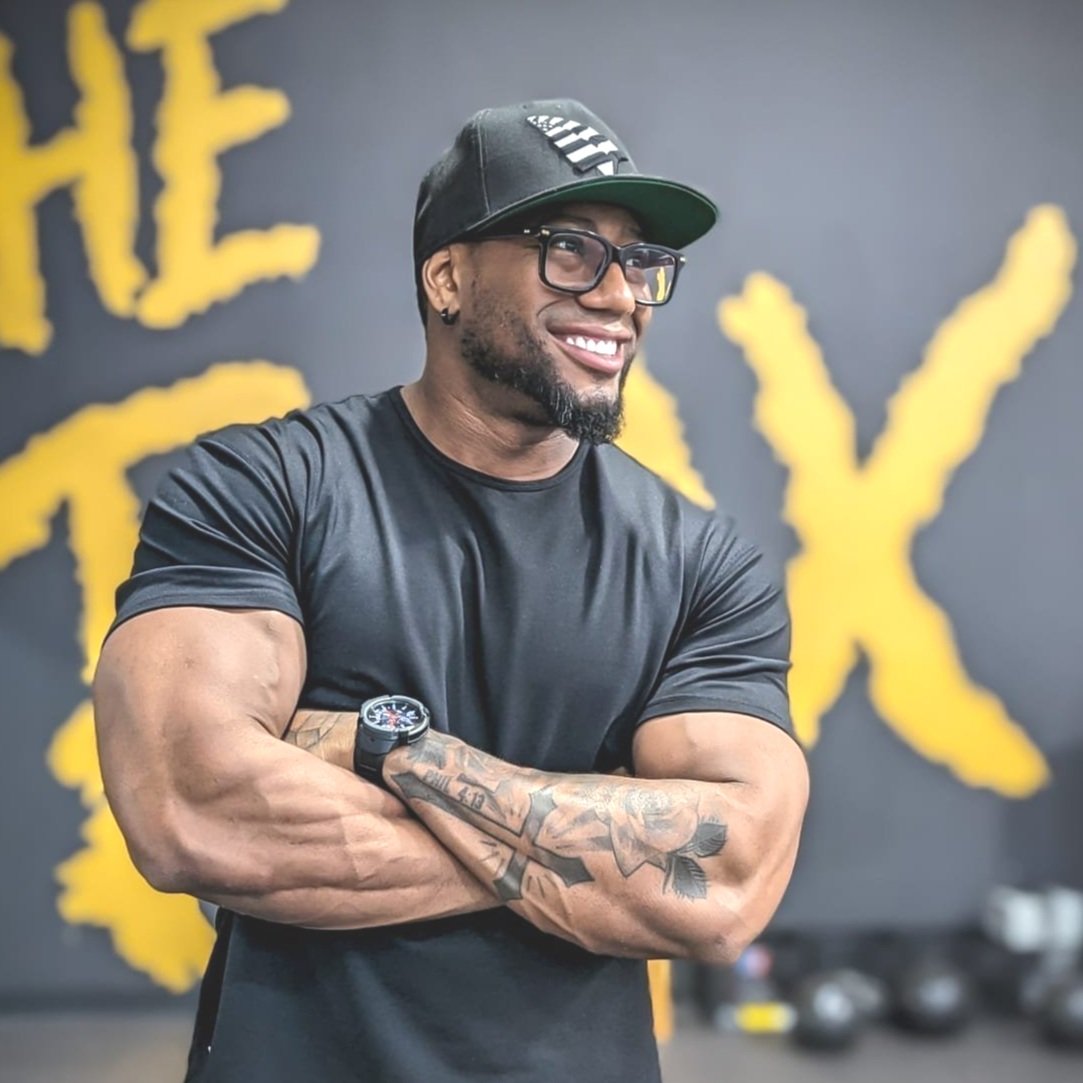 Smiling man with muscular arms, tattoos, glasses, black baseball cap, black T-shirt, standing in gym with yellow and black logo in background.