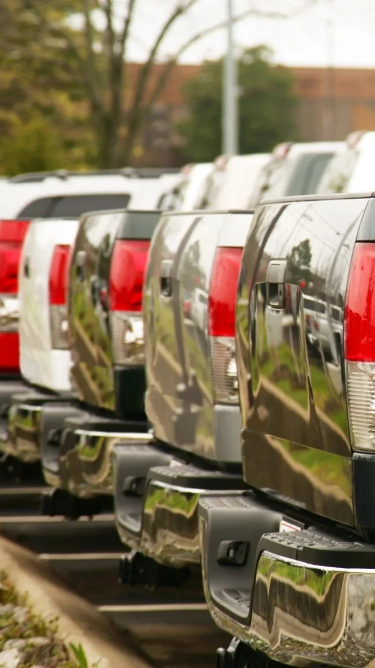 Multiple pickup trucks lined up in a row in a parking lot, with reflected trees and sky on their shiny chrome backs.