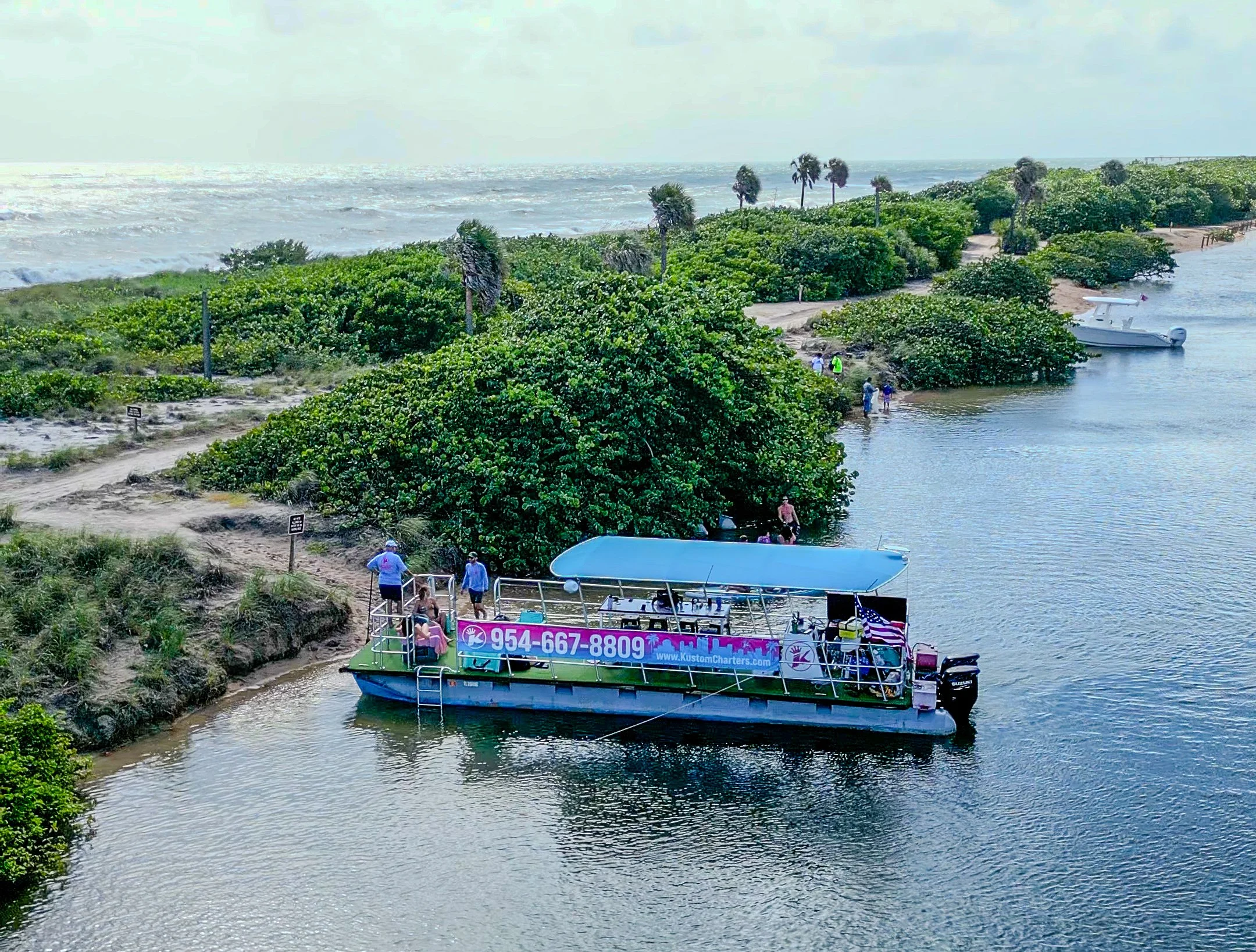 A boat docked at a small river inlet, with a sign displaying a phone number and website, surrounded by lush green bushes and palm trees, and a sandy beach with people walking in the distance, near the ocean.