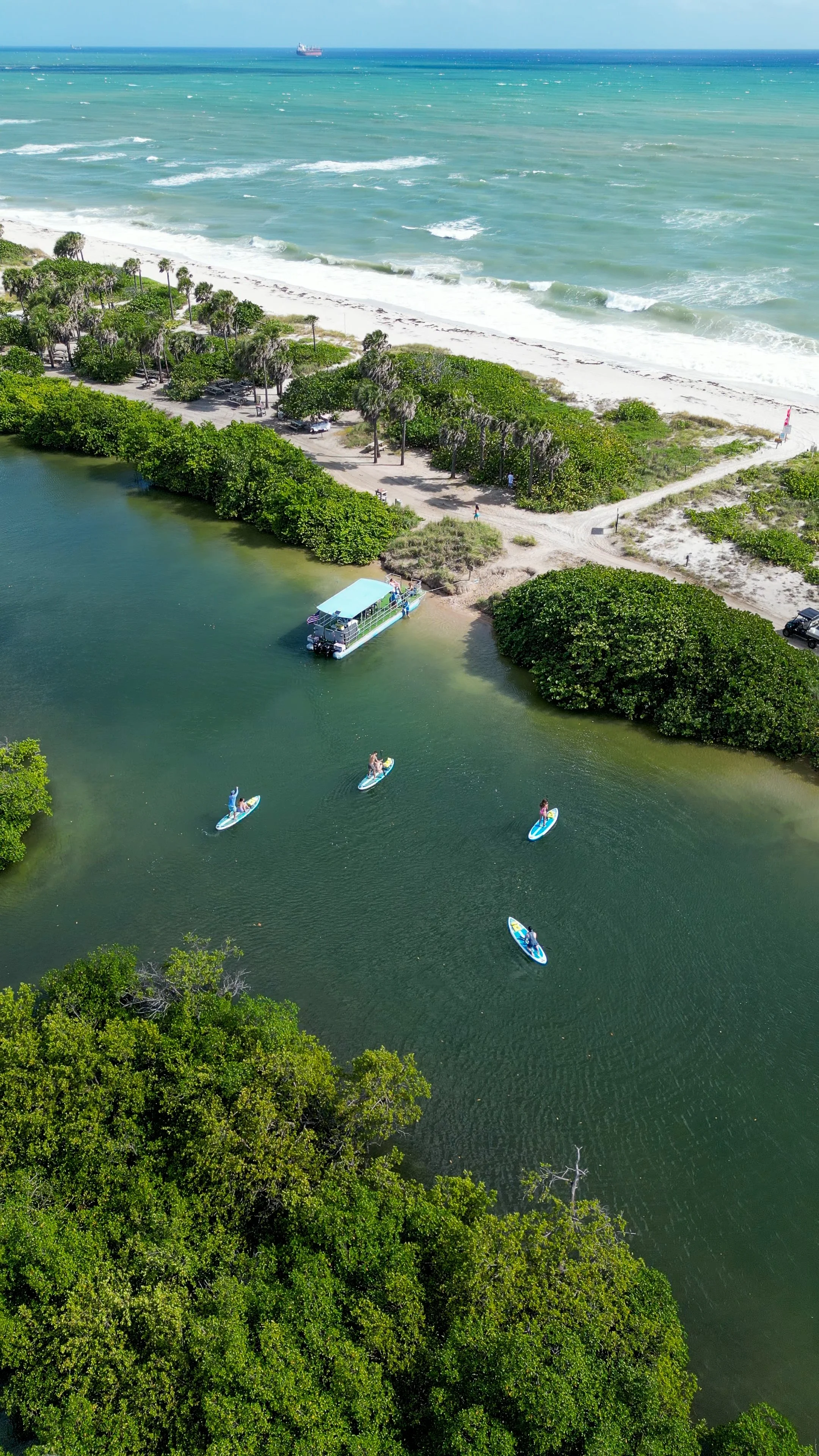 An aerial view of a coastal area featuring a beach with white sand, a green waterway with paddleboarders, paddleboats, lush greenery, and a boat along the shoreline.