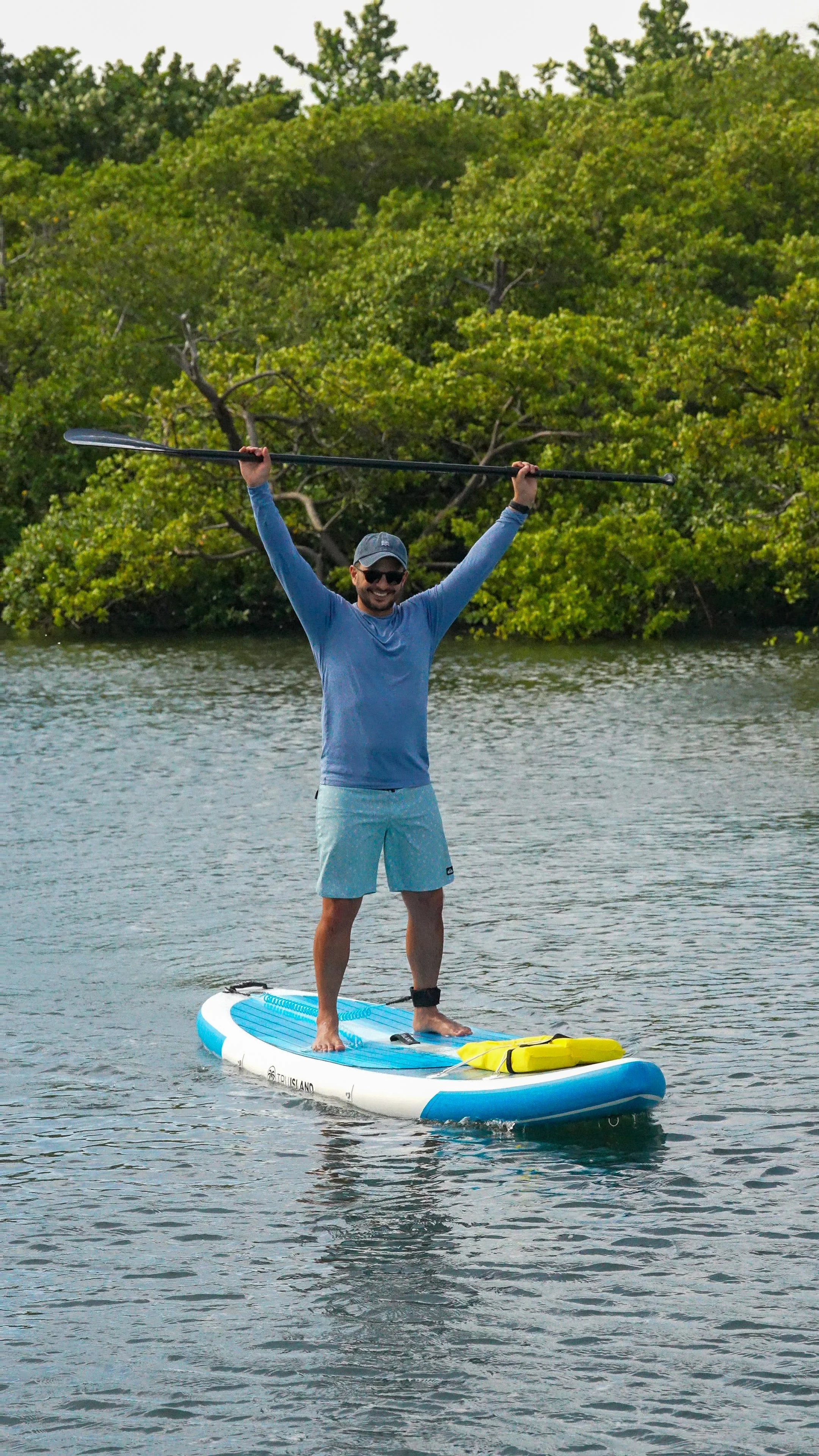 A man standing on a paddleboard in the water, holding a paddle above his head, smiling, wearing sunglasses, a gray cap, a blue long-sleeve shirt, and light blue shorts.