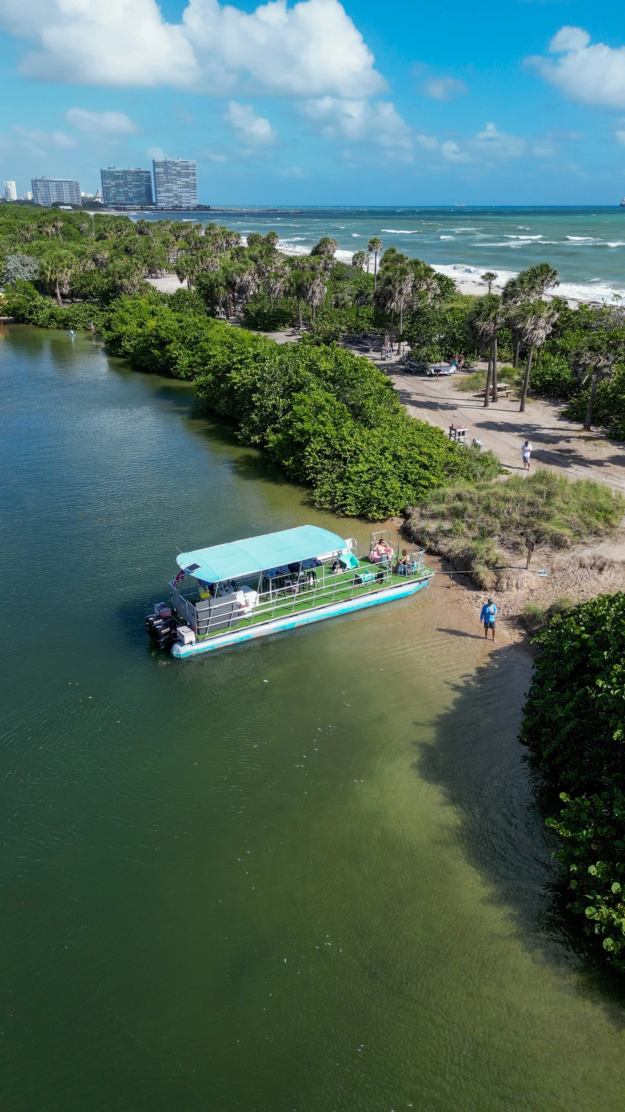 A boat docked along a river near a sandy beach with palm trees, with the ocean and city buildings in the background.