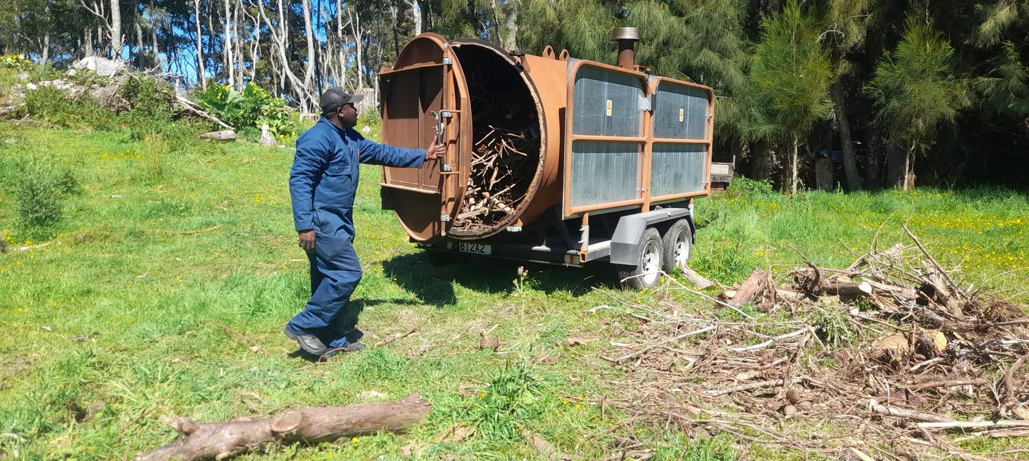 A man in a blue jumpsuit and black cap operating a wood chipper, feeding branches into it in a grassy, wooded outdoor area.