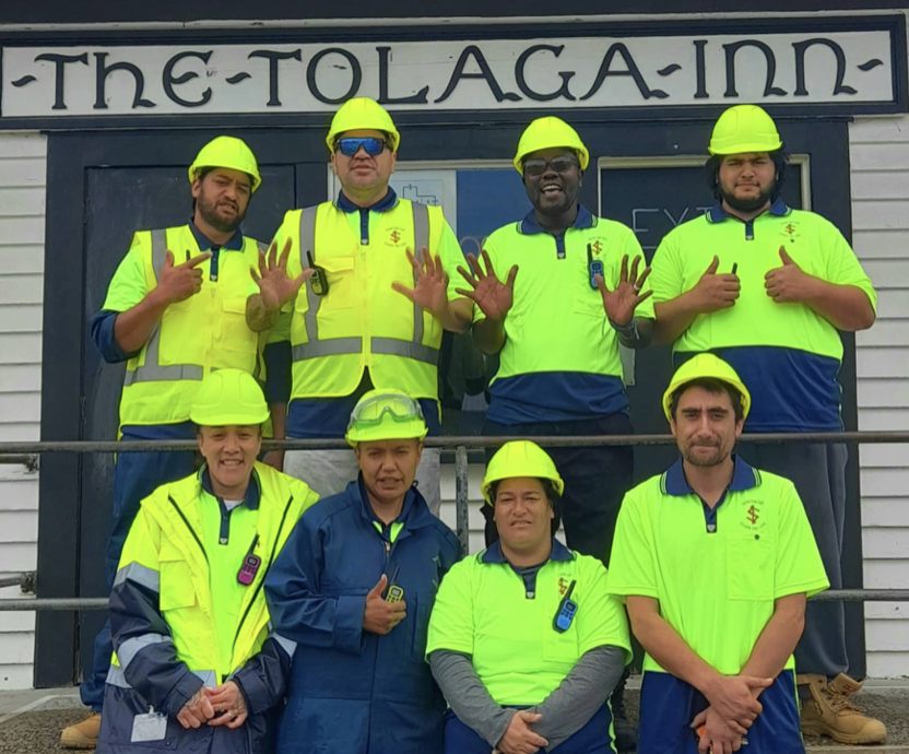 Group of nine workers in safety vests and helmets standing in front of The Tolaga Inn. Some are smiling, and a few are making hand gestures with a building in the background.