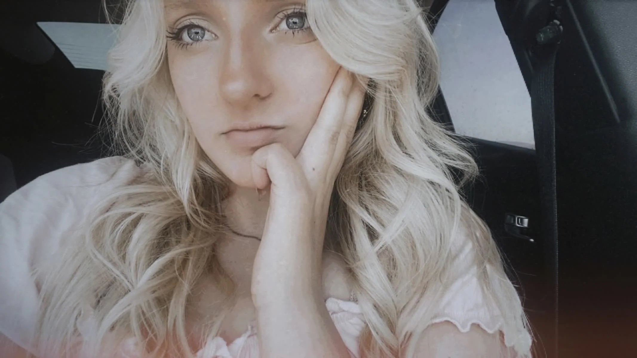 A woman with blonde wavy hair and blue eyes resting her face on her hand in a car.