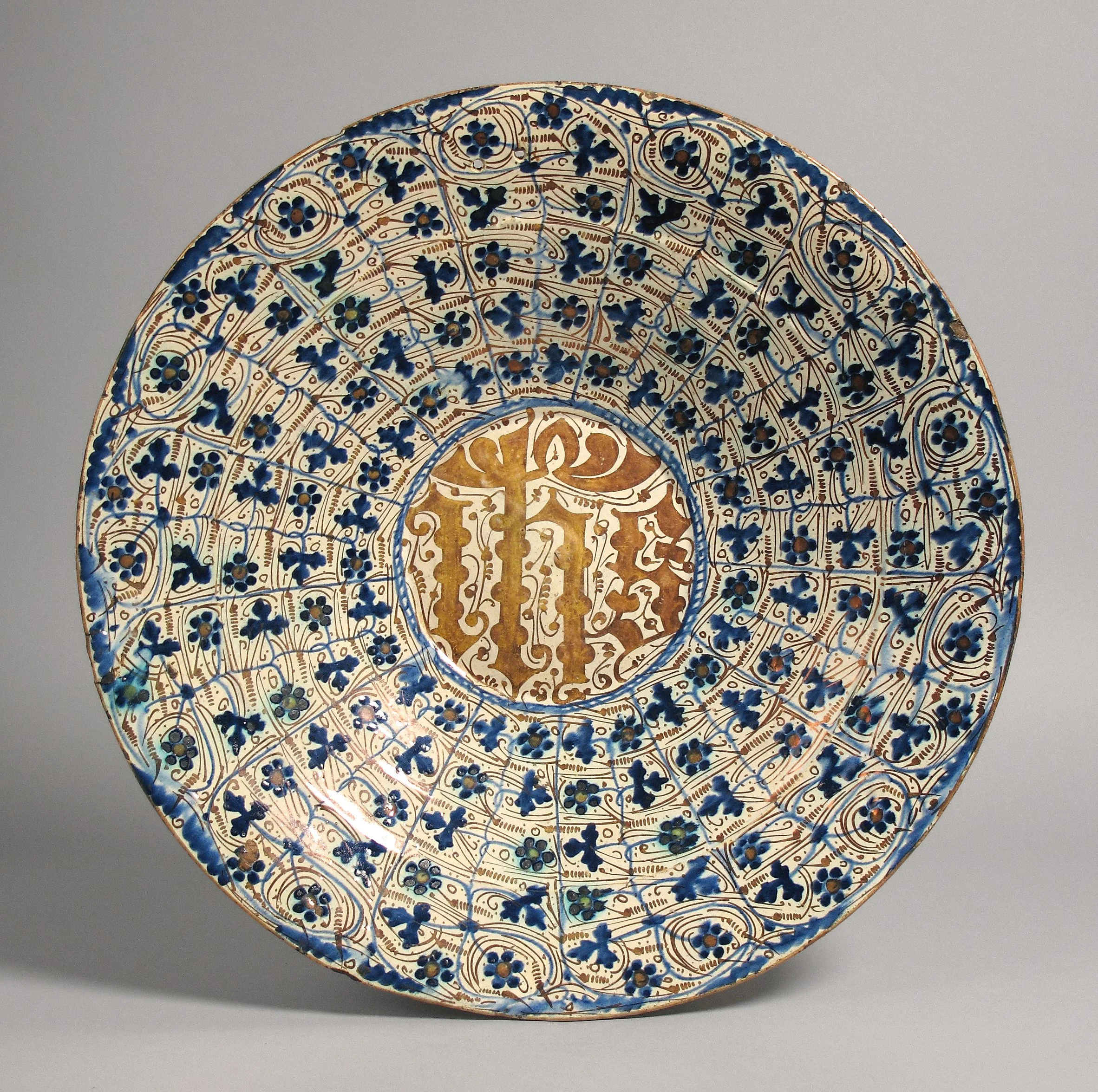 A decorative ceramic plate with intricate gold and blue floral and geometric patterns, displayed against a plain gray background.