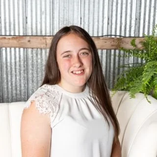Smiling woman sitting on a white sofa, against a backdrop of corrugated metal and wood, with a fern plant nearby, wearing a white blouse with lace sleeves.