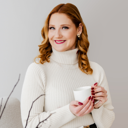 Smiling person with long, wavy blonde hair in a light-colored blouse, standing against a dark striped background.