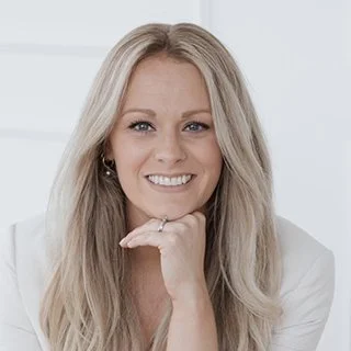 Portrait of a smiling woman with long blonde hair, wearing a light-colored outfit, posing with her hand under her chin.