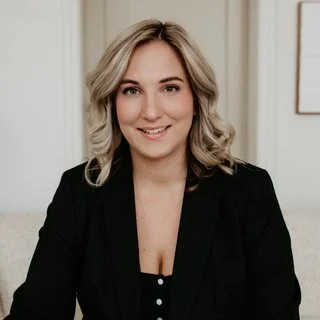 Smiling person with shoulder-length blonde hair wearing a black blazer, seated indoors.