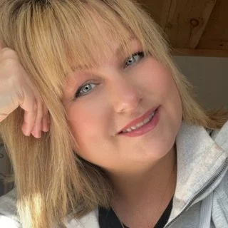 Smiling woman with blonde hair, wearing a light jacket, resting her head on her hand, in a well-lit indoor setting.