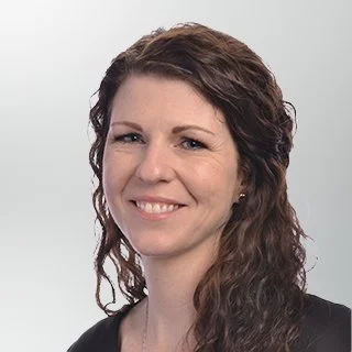 A woman with long wavy brown hair smiling, wearing a black top against a plain light background.
