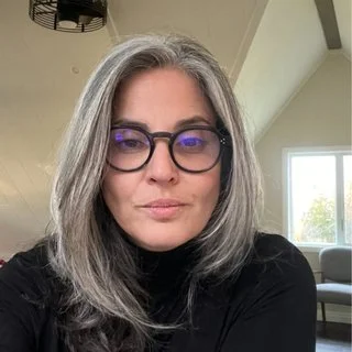 A person with long gray hair and glasses sitting in a well-lit room.