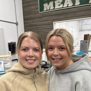 Two women smiling in a store with a sign that reads 'MEAT' in the background.