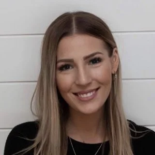 Smiling woman with long blonde hair wearing a black top, standing against a white wall background.