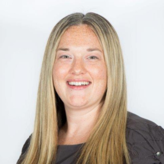 Smiling person with long blonde hair wearing a dark top against a plain background.