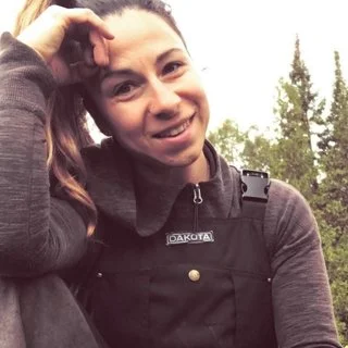 Person smiling outdoors wearing dark overalls, with trees in the background.