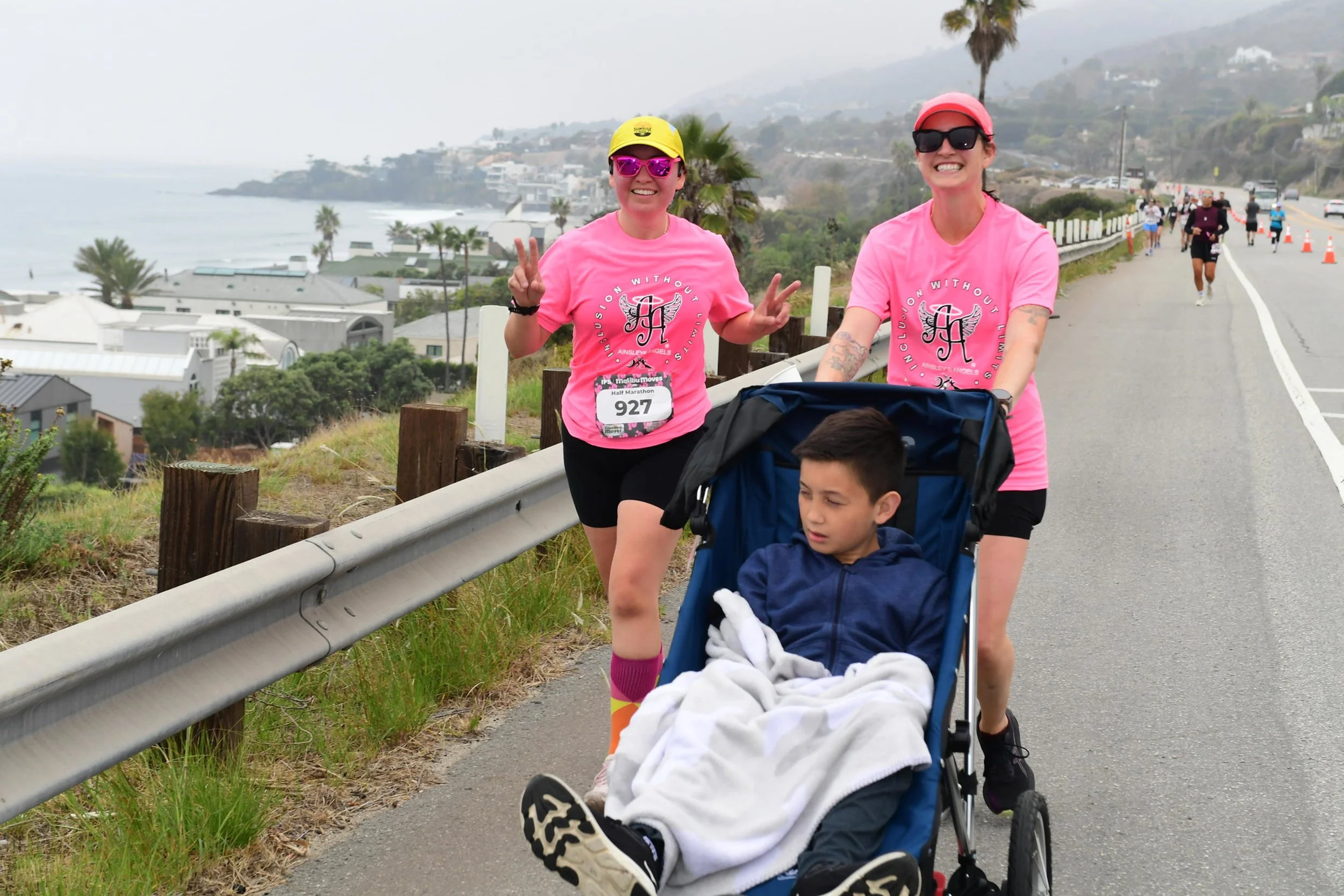 Marce and Leah in matching pink Malibu Moves shirts run along PCH pushing Marce's son in a race chair, with the Malibu coastline and ocean visible behind them. Marce flashes a peace sign and both are smiling.