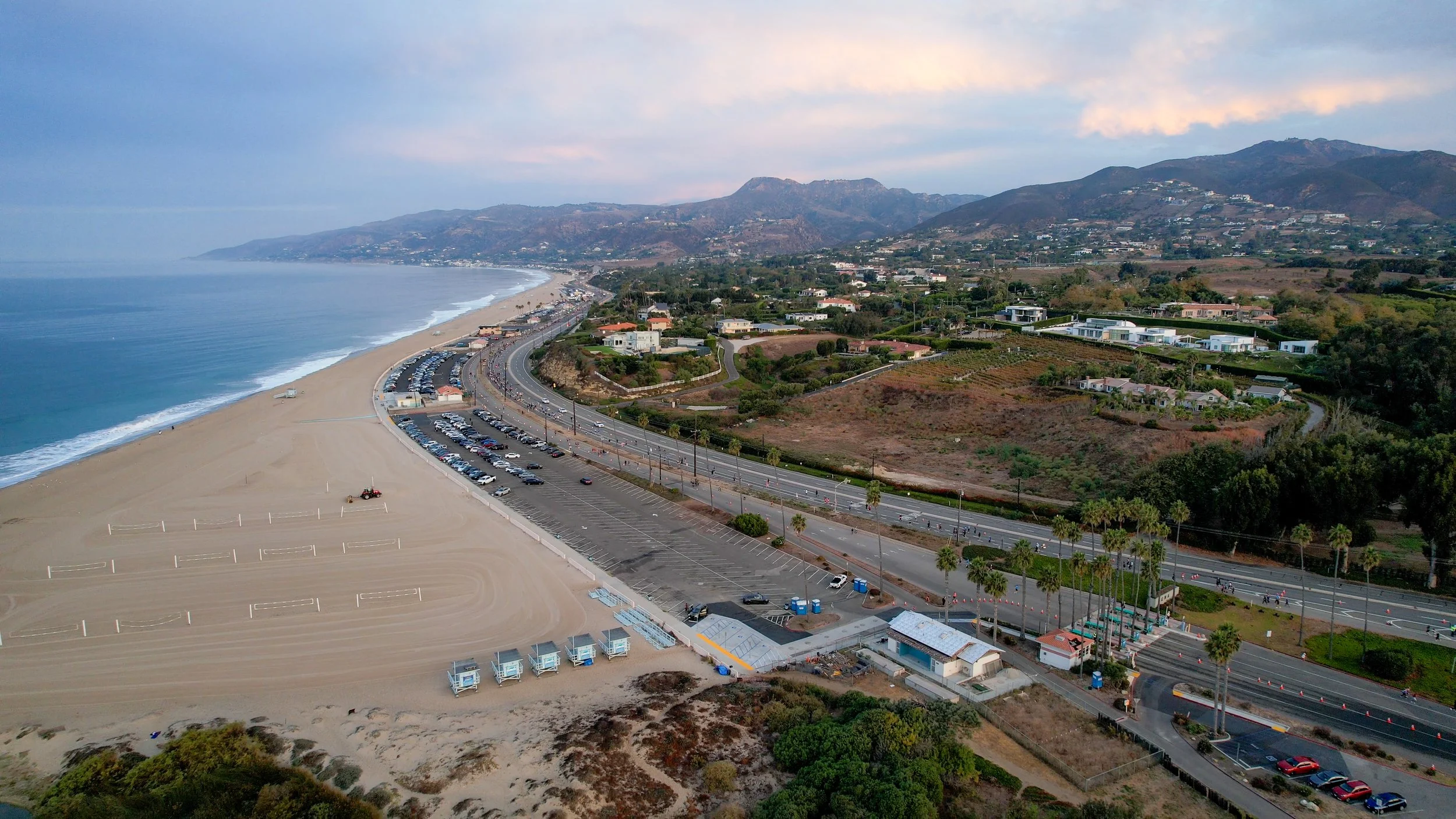 Aerial view of Zuma Beach at sunrise on race morning, showing the beach parking lots along the sand and runners on Pacific Coast Highway below the Santa Monica Mountains.