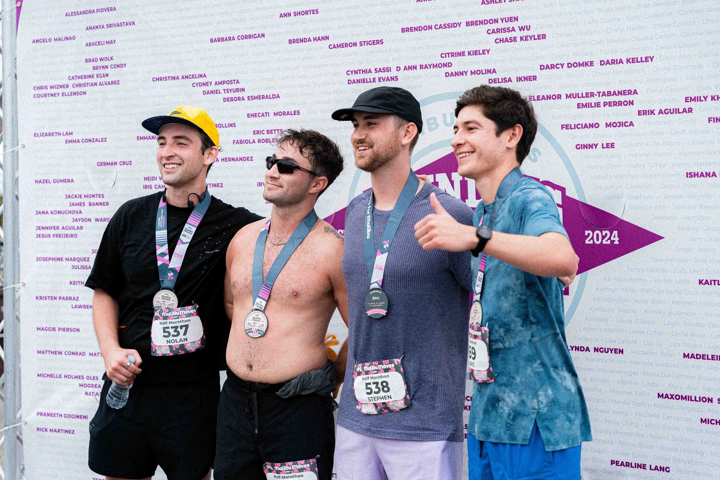 Four Malibu Moves runners stand together wearing their finisher medals in front of the Founding Movers 2024 wall, one giving a thumbs up.