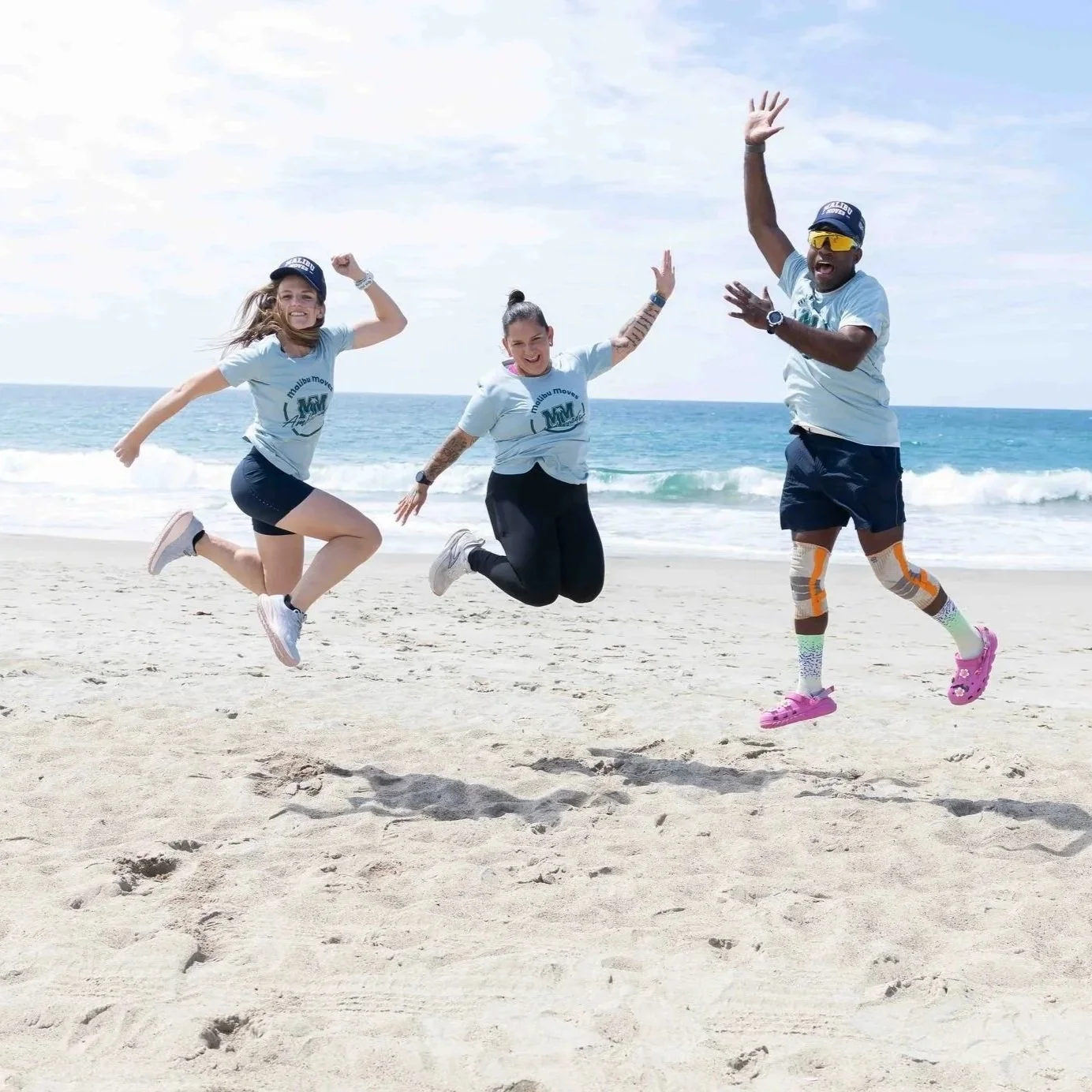 Three people jumping in the air on a sandy beach with the ocean and sky in the background. They are smiling, wearing light blue T-shirts, and sneakers. One person is wearing colorful leg sleeves and pink sandals, another has tattoos and black pants, and the third has a cap and sunglasses.