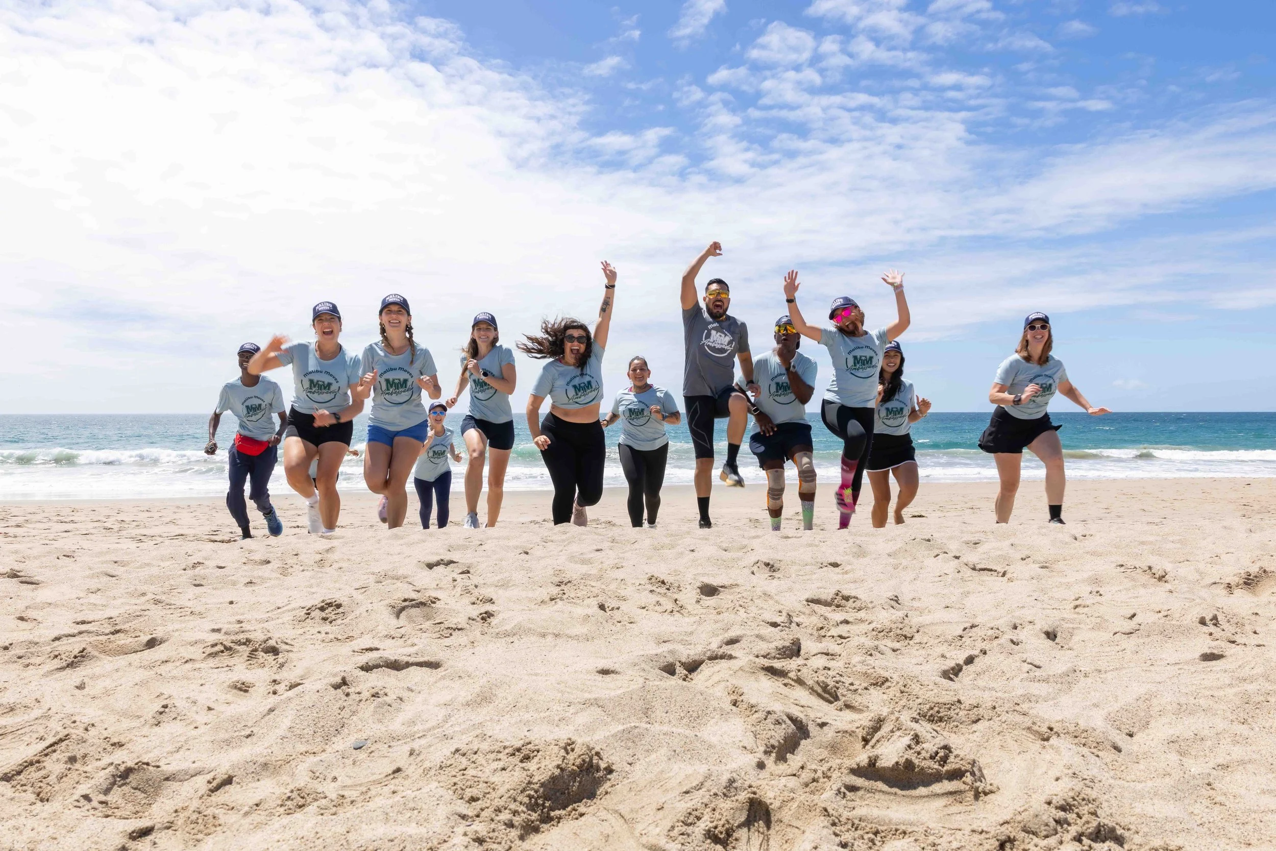 A group of Malibu Moves Ambassadors running and jumping on a sandy beach near the ocean under a partly cloudy sky. They are wearing Malibu Moves' swag.
