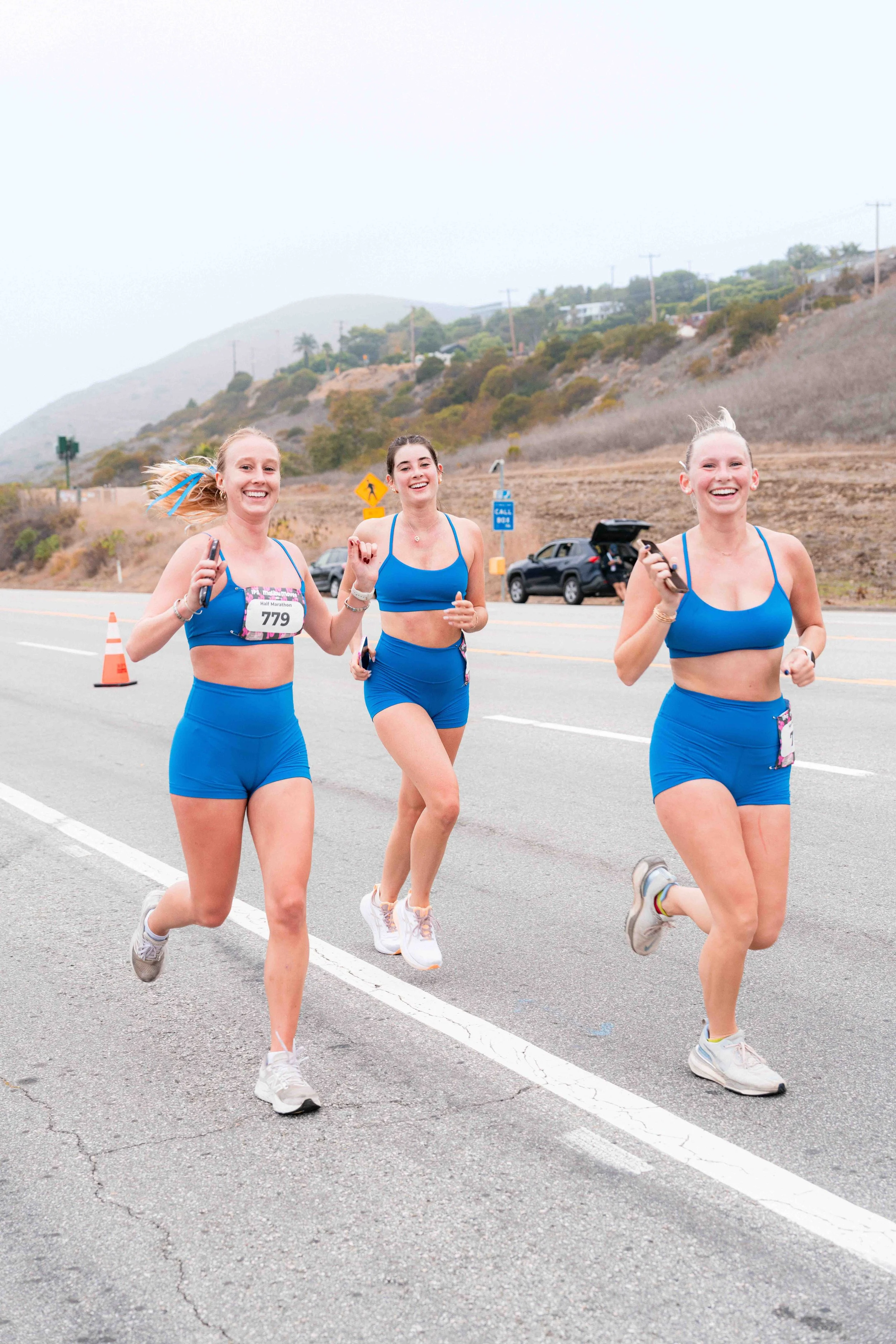 Three women running Malibu's official road race, all wearing blue athletic outfits and smiling. The background features hills, trees, a car with an open trunk, and some road signs, under an overcast sky.