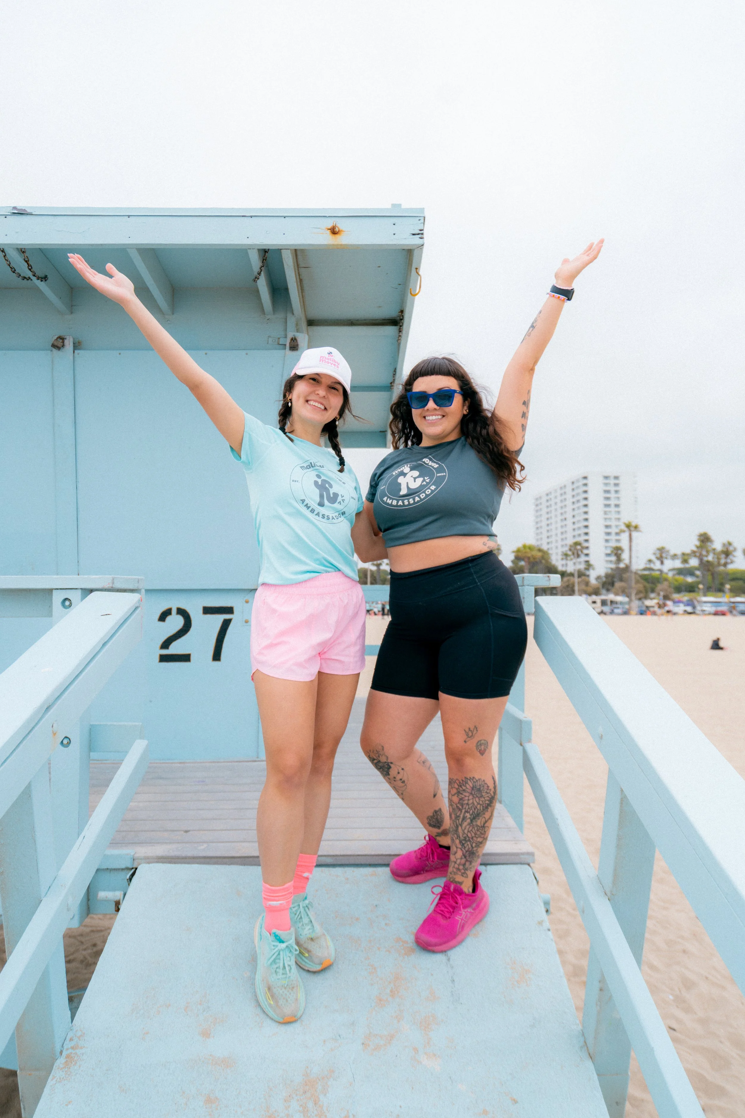 Two Malibu Moves ambassadors posing with arms raised on a lifeguard tower at Zuma Beach in Malibu.