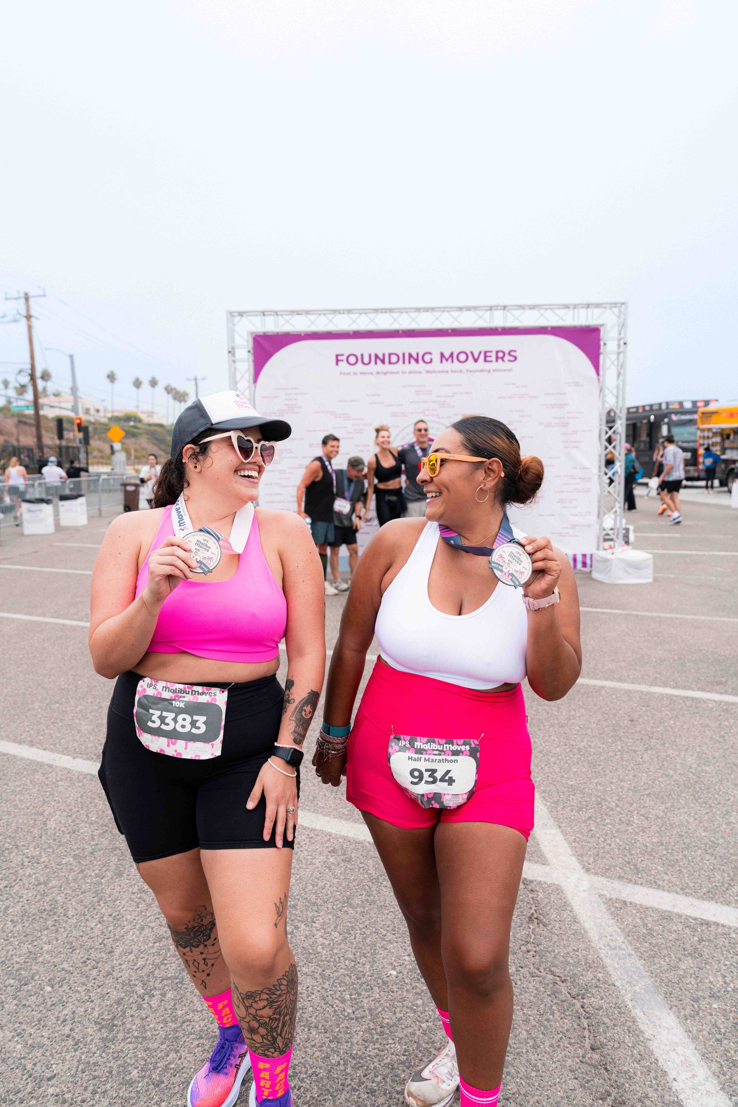 Two women walking and smiling after finishing Malibu Moves, holding medals in their hands, with a purple-themed event banner reading "Founding Movers" in the background at an outdoor race event.