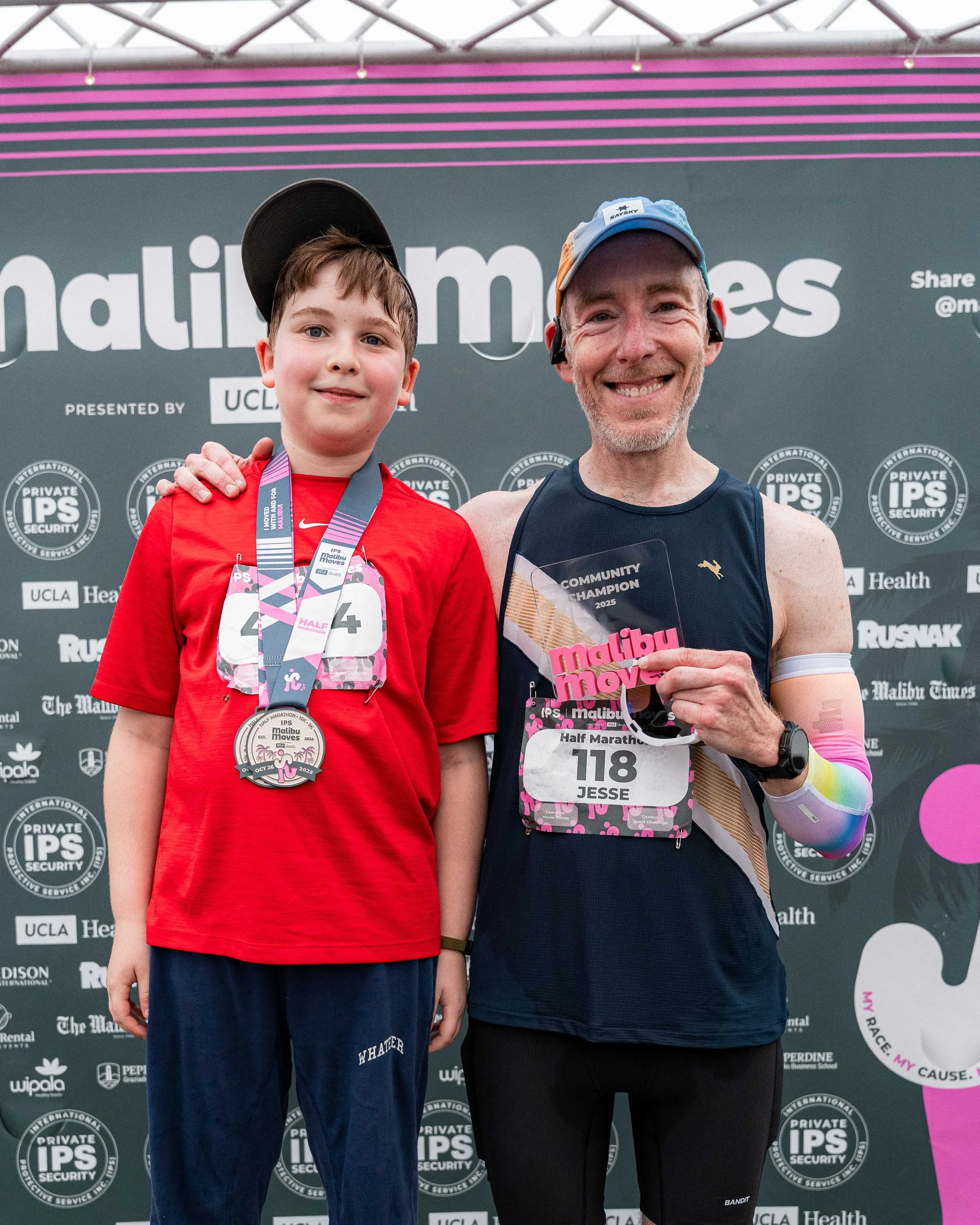 A smiling father and son wearing their Malibu Moves finisher medals and holding a 'Community Champion' award for his fundraising effort in front of the race backdrop.