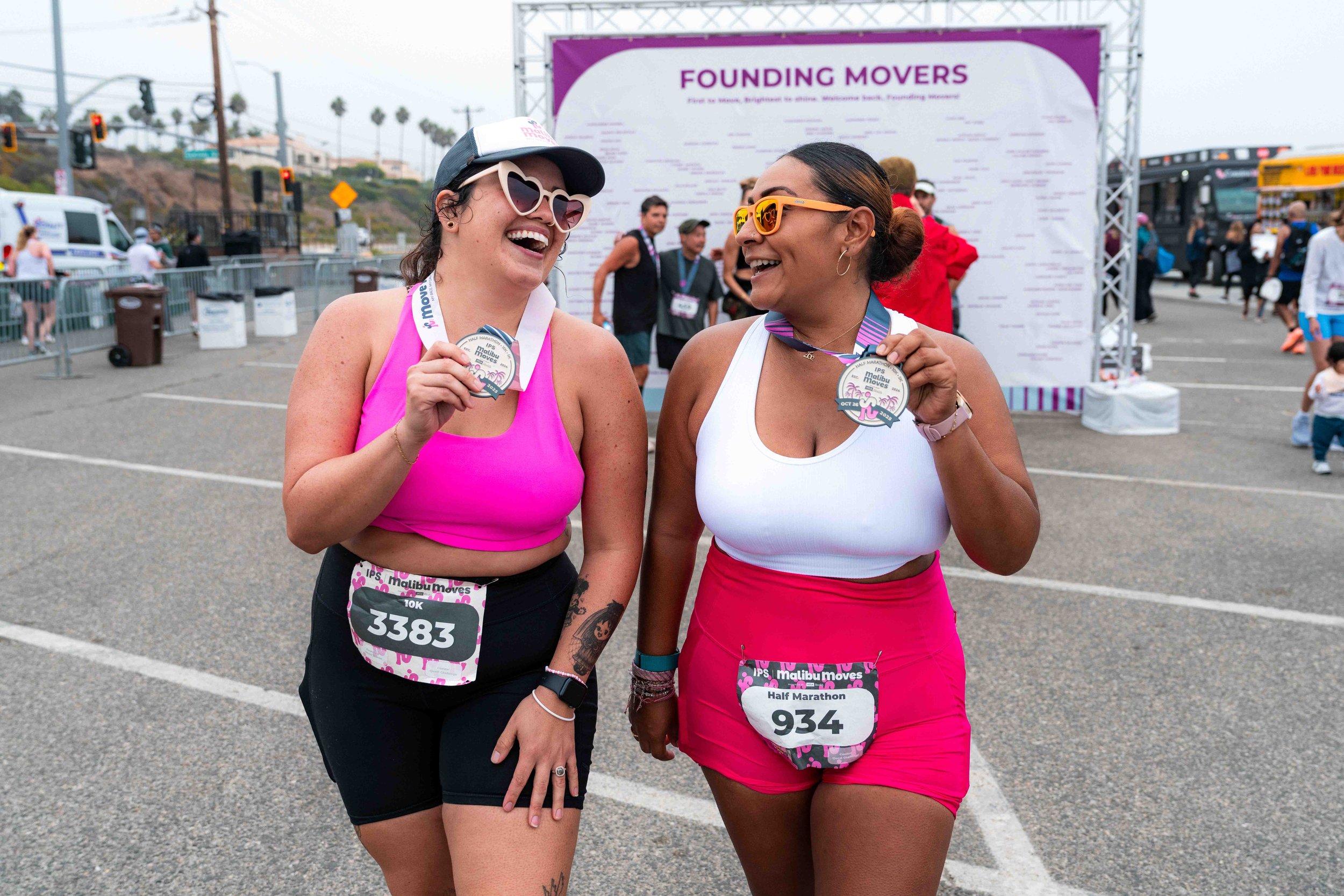 Two Malibu Moves participants smiling and holding their finisher medals after crossing the finish line at Zuma Beach.