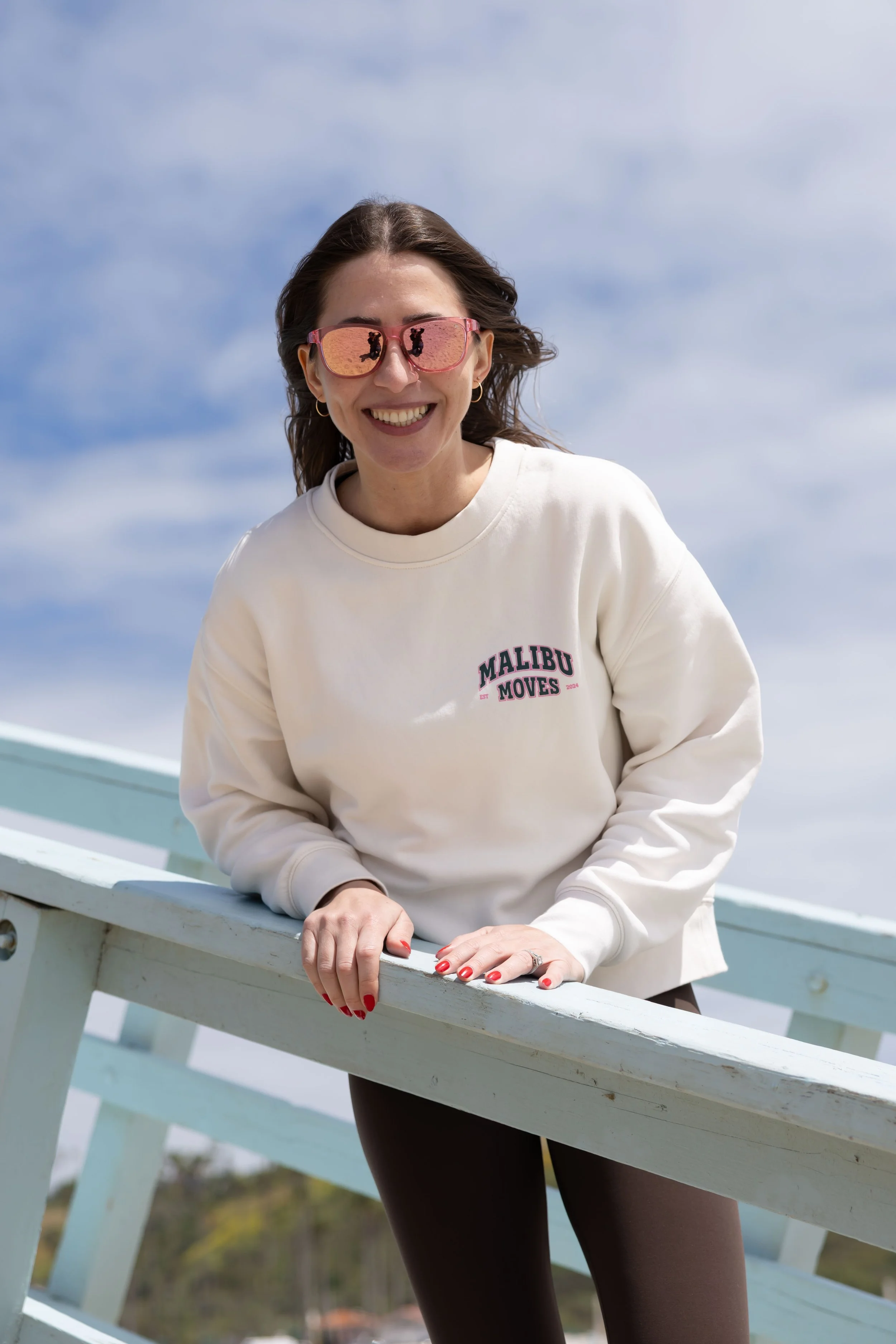 Erica Segel, founder of Malibu Moves, smiles while standing on the beach in a light pink sweatshirt, with the ocean in the background.