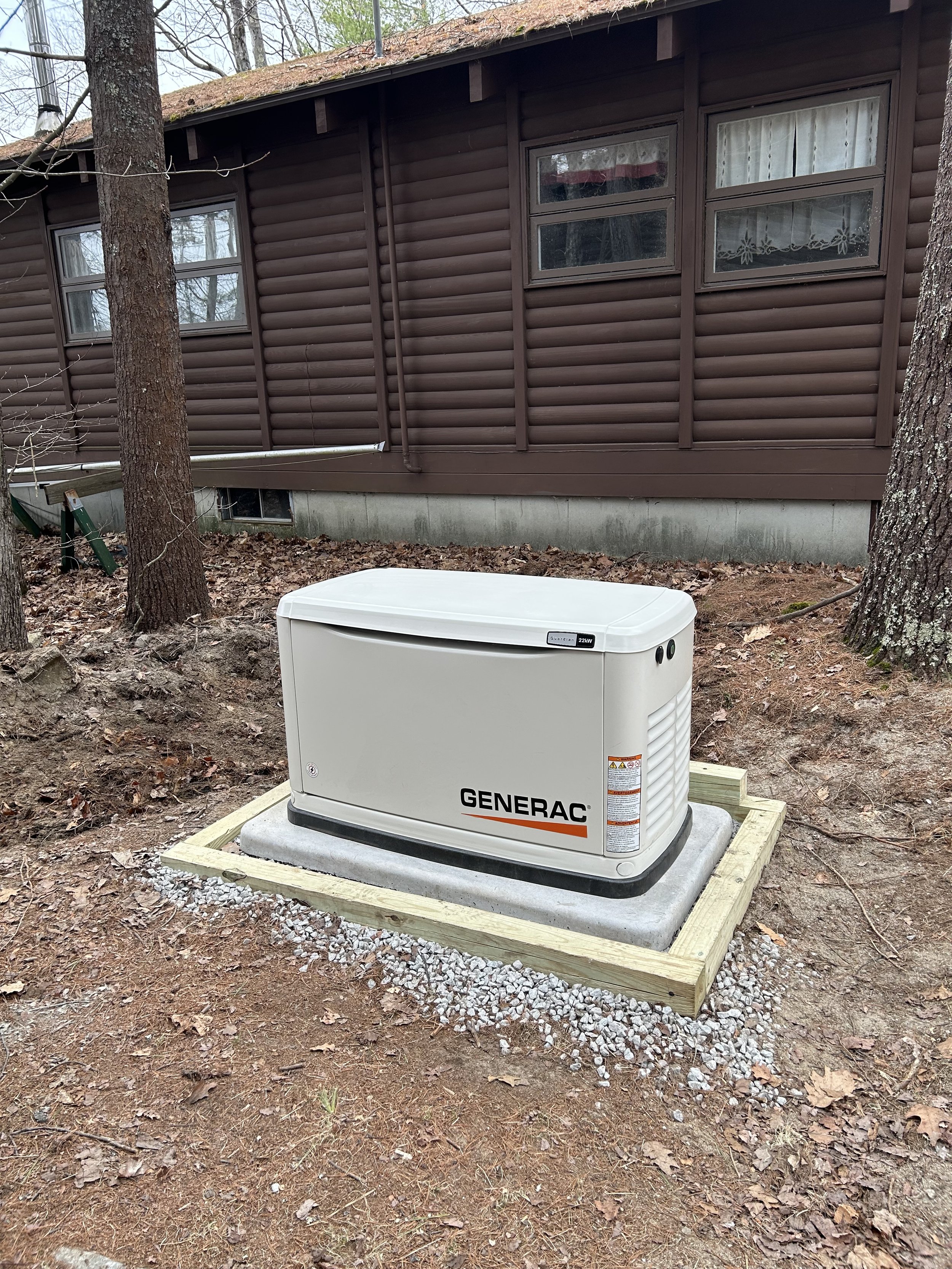 A Generac home backup generator installed outdoors on a concrete slab, surrounded by a wooden frame and gravel, near a brown house with trees in the background.