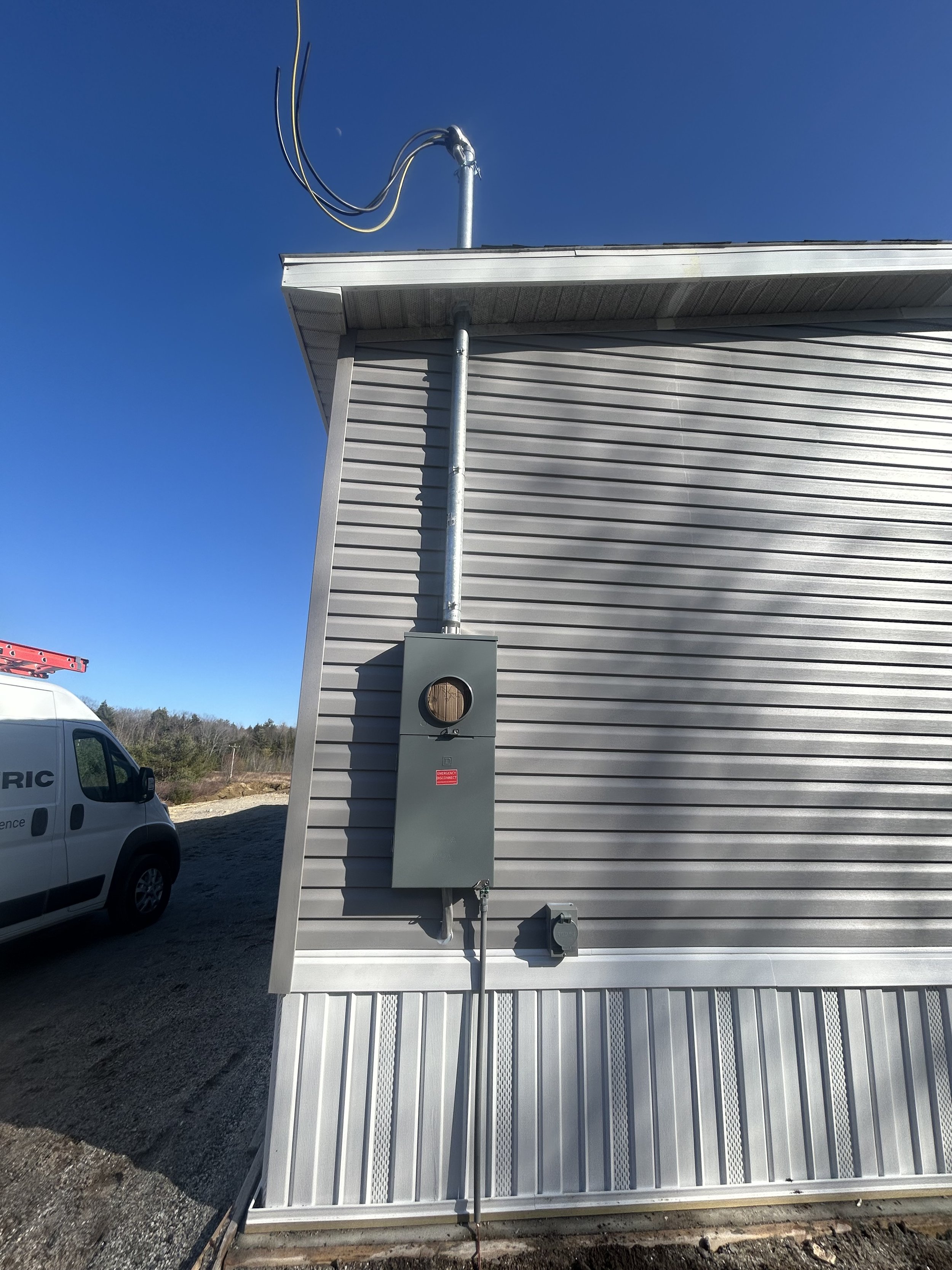 Exterior view of a gray vinyl-sided building with an electric meter box and conduit pipe on the wall, a utility van nearby, and a clear blue sky.