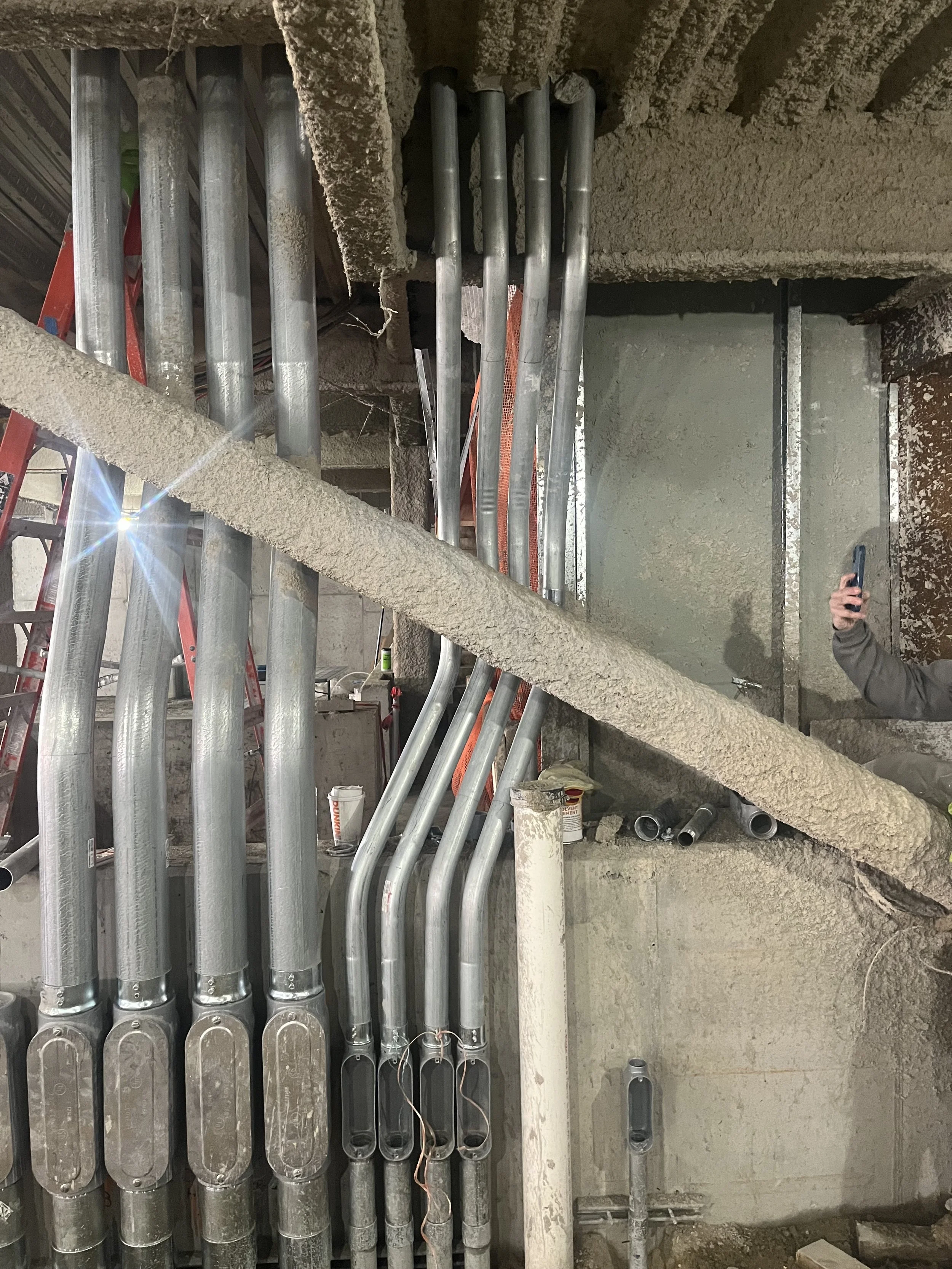 Electrical conduits running vertically along a rough concrete wall in an industrial setting. A person holding a smartphone is partially visible on the right side. Red ladder and construction materials are seen in the background.