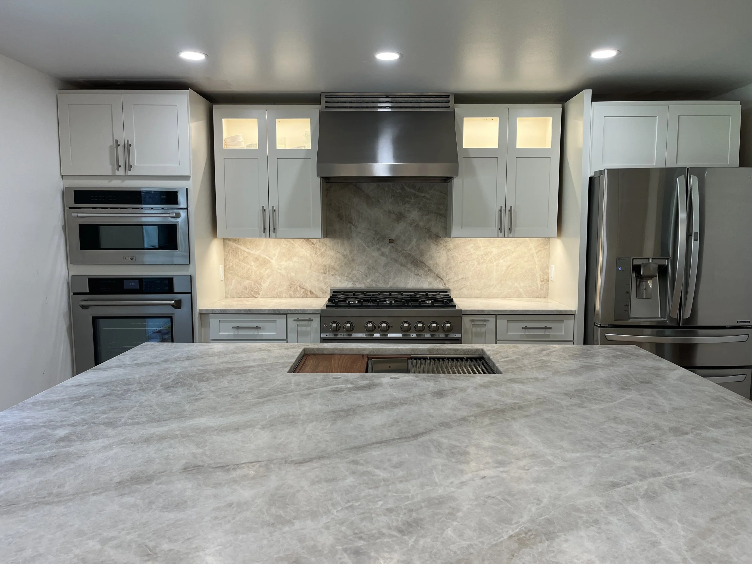 Modern kitchen with white cabinets, stainless steel appliances including a refrigerator, oven, and microwave, gray marble backsplash, and a marble countertop island.