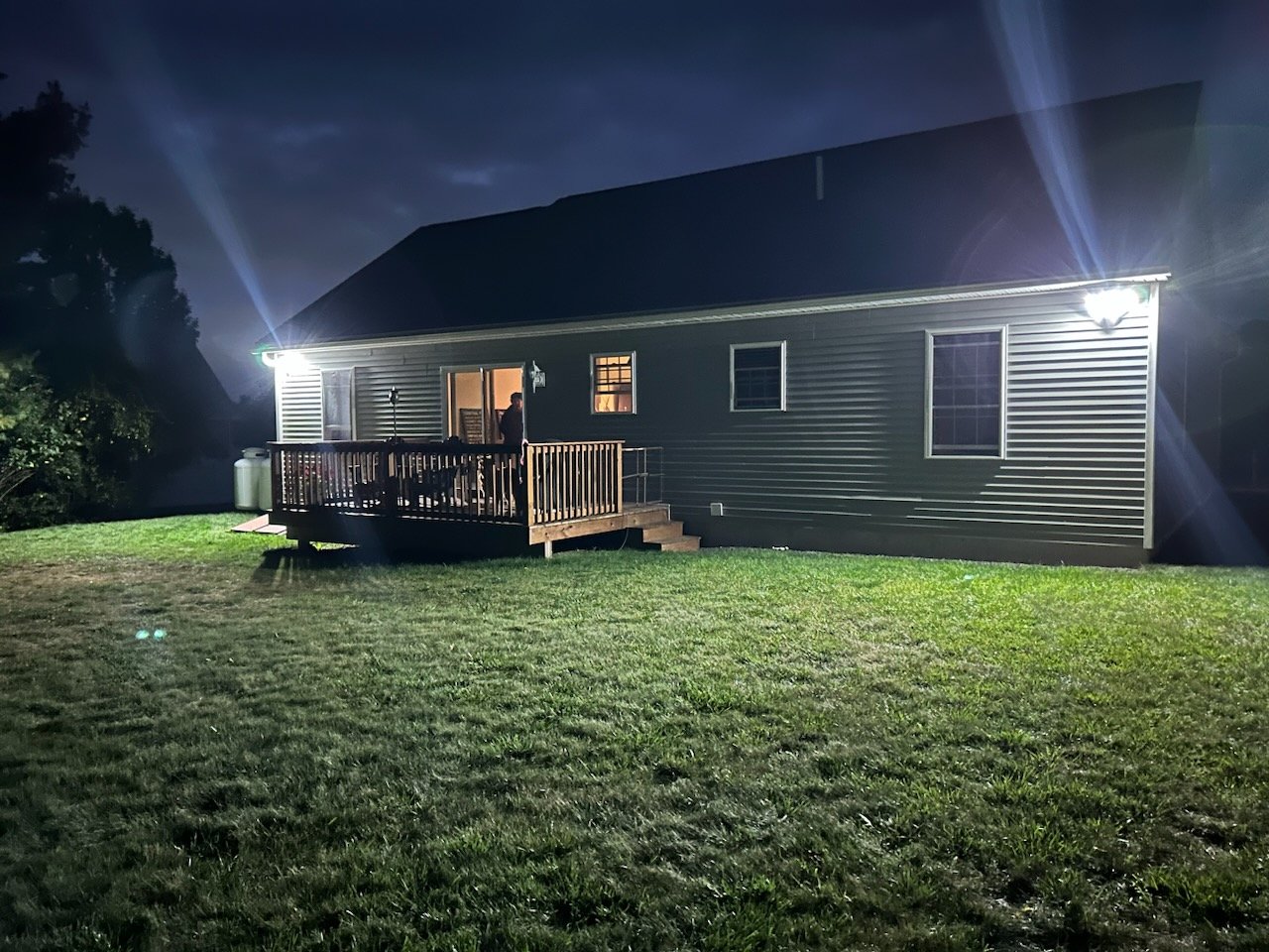 Backyard view of a house with lights illuminating a wooden deck at night. The lawn is visible in the foreground.