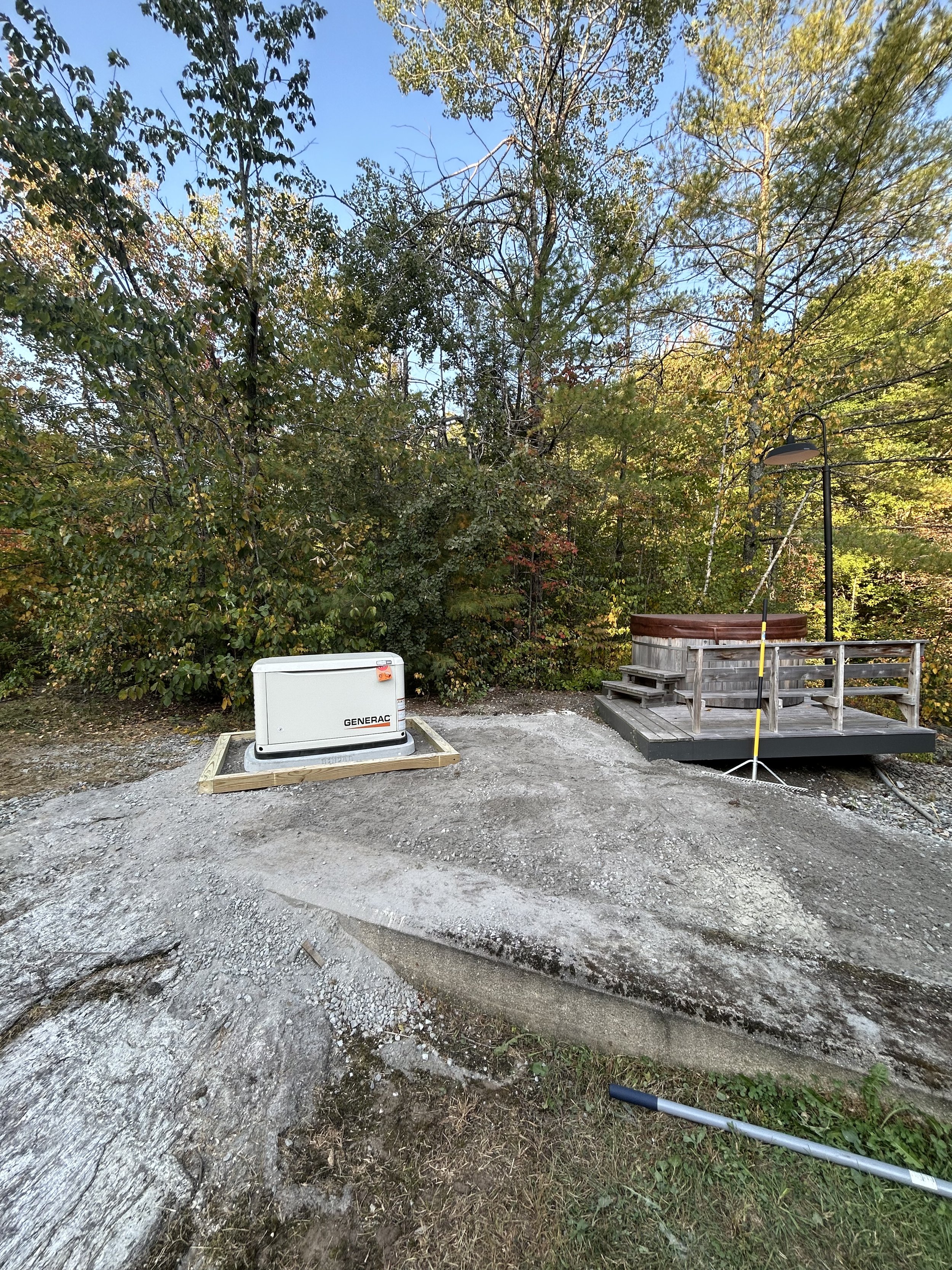 Outdoor scene with a Generac generator on a concrete slab next to a wooden platform surrounded by trees.