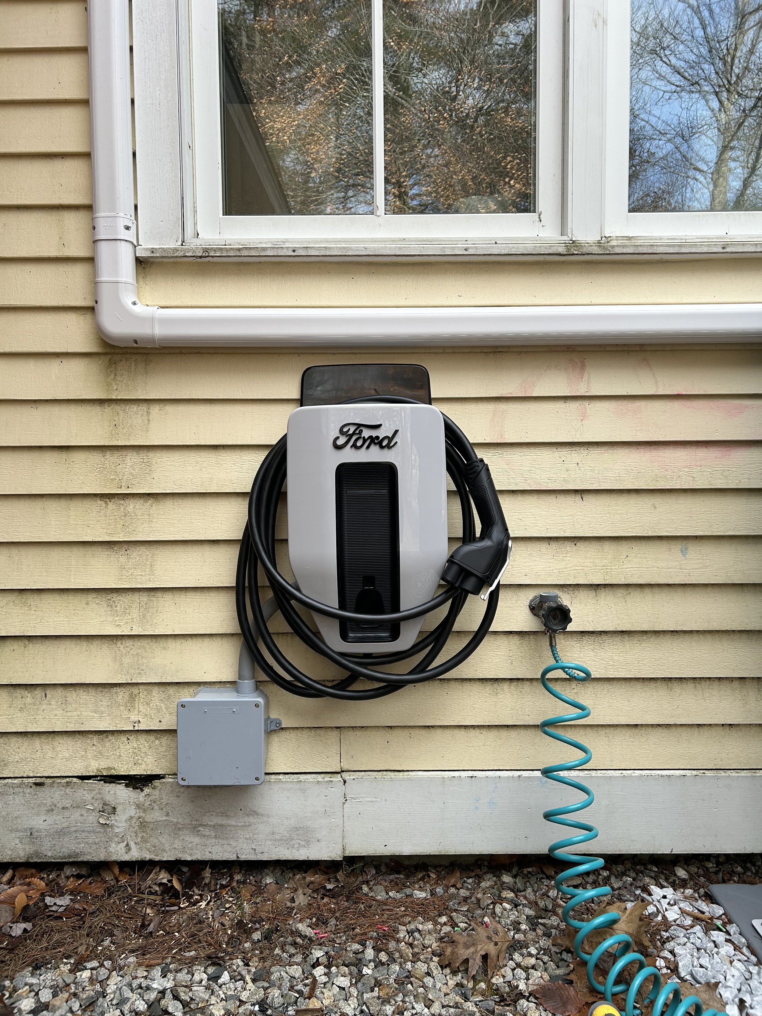 Ford electric vehicle charging station mounted on house exterior wall, with a coiled hose nearby.