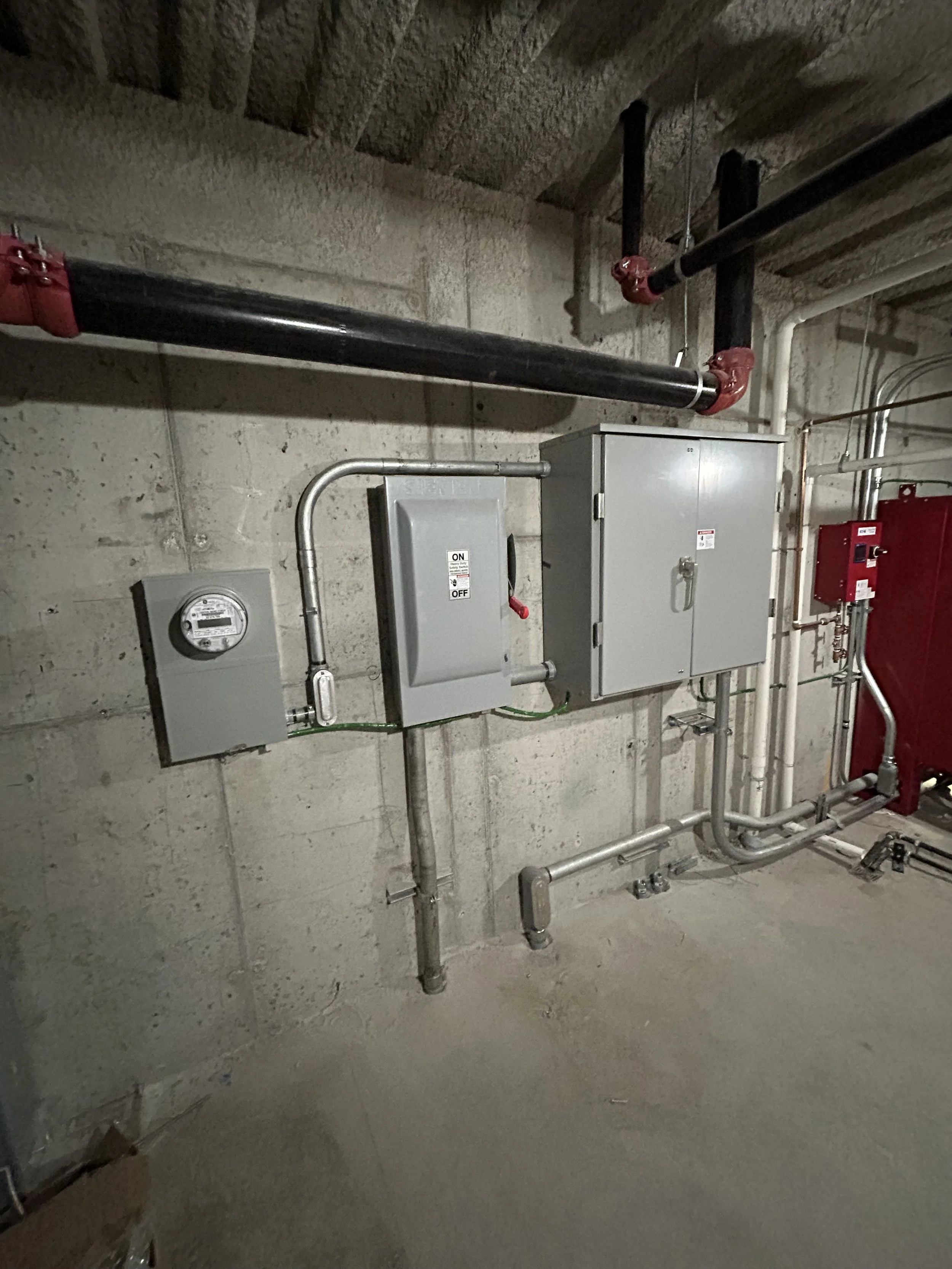 Industrial electrical control panel with pipes and gray metal boxes mounted on a concrete wall in a utility room.