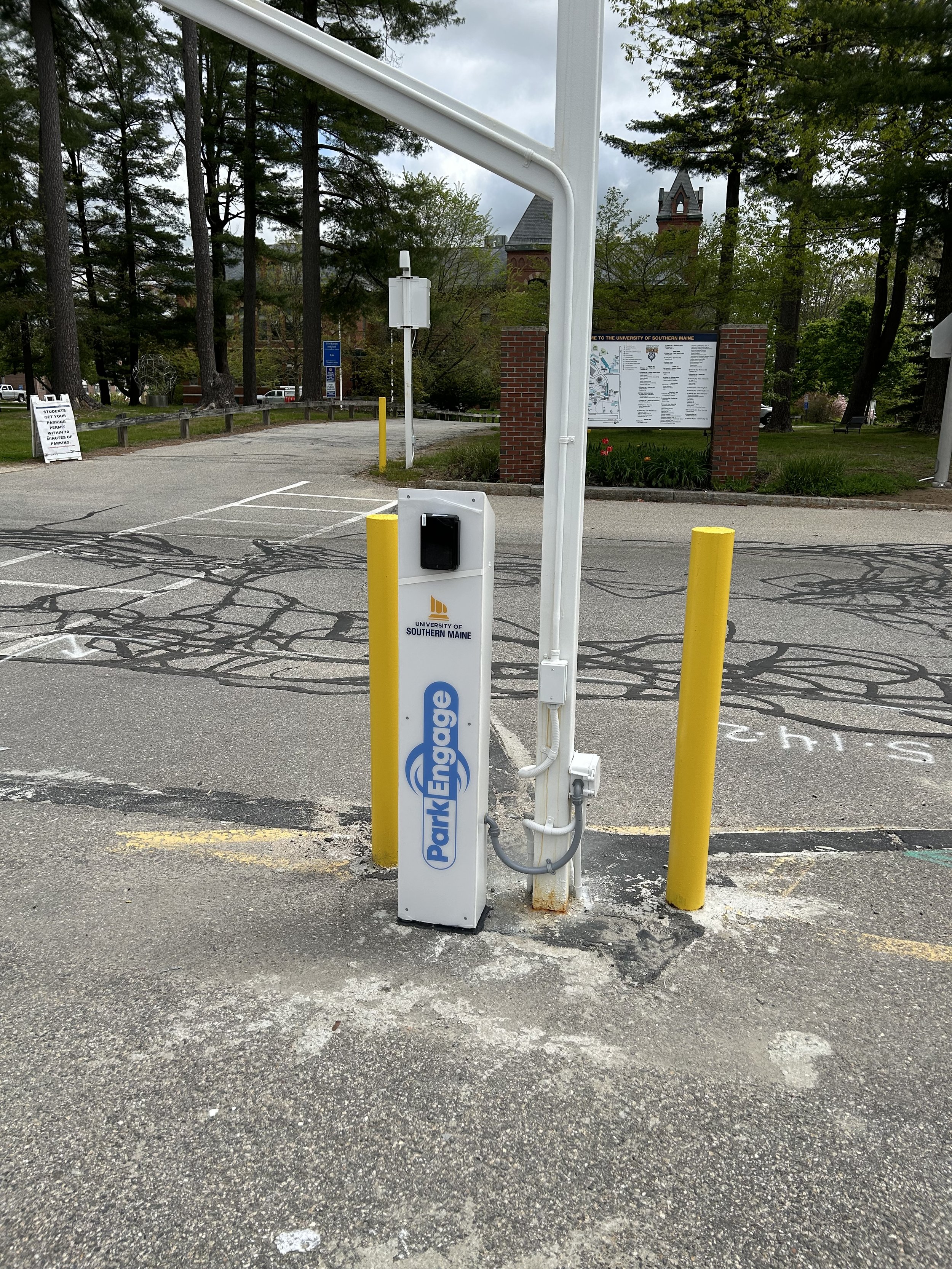 Parking lot entrance with ParkEngage ticket kiosk and yellow posts. Trees and a building in the background.