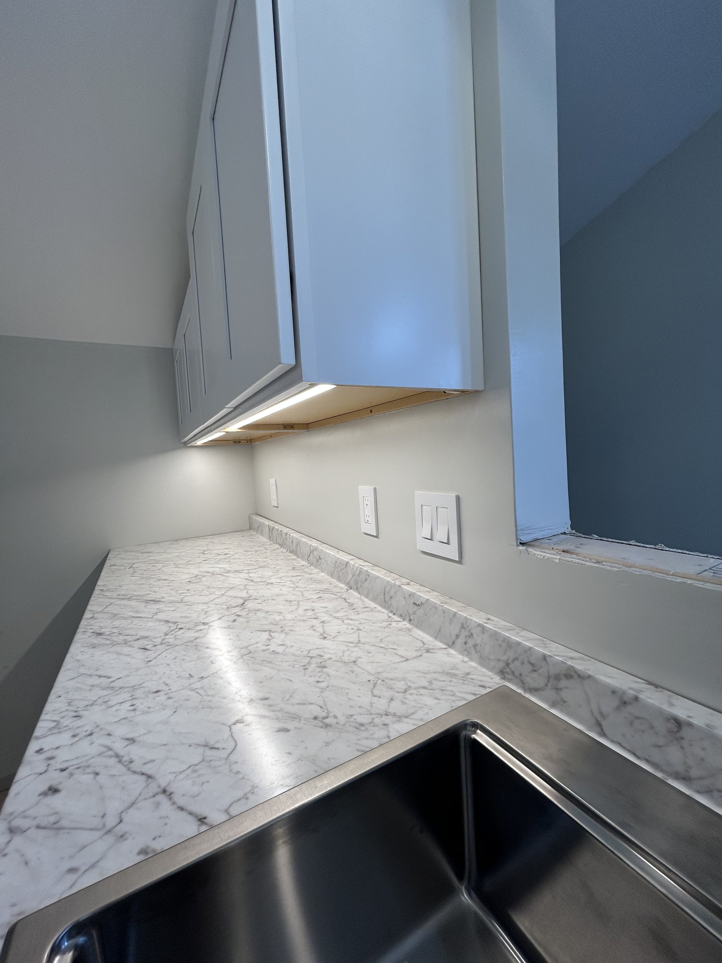 Modern kitchen with marble countertop, stainless steel sink, white cabinets, and wall light switches.