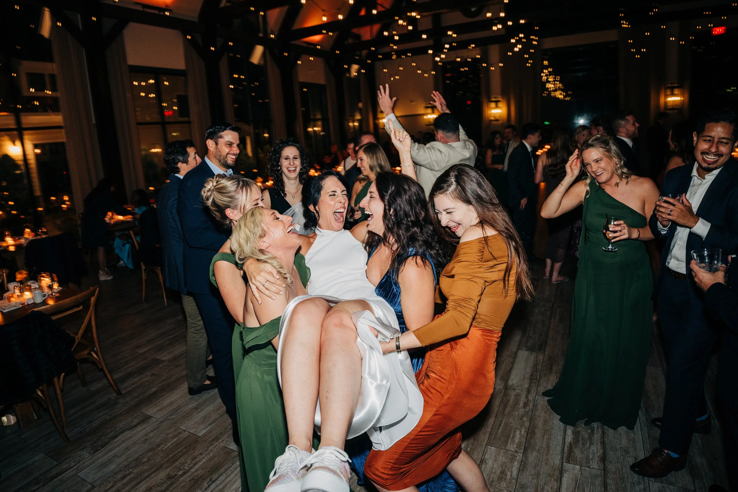 Group of people dancing and celebrating at a party or wedding reception with string lights and decorated tables in the background.