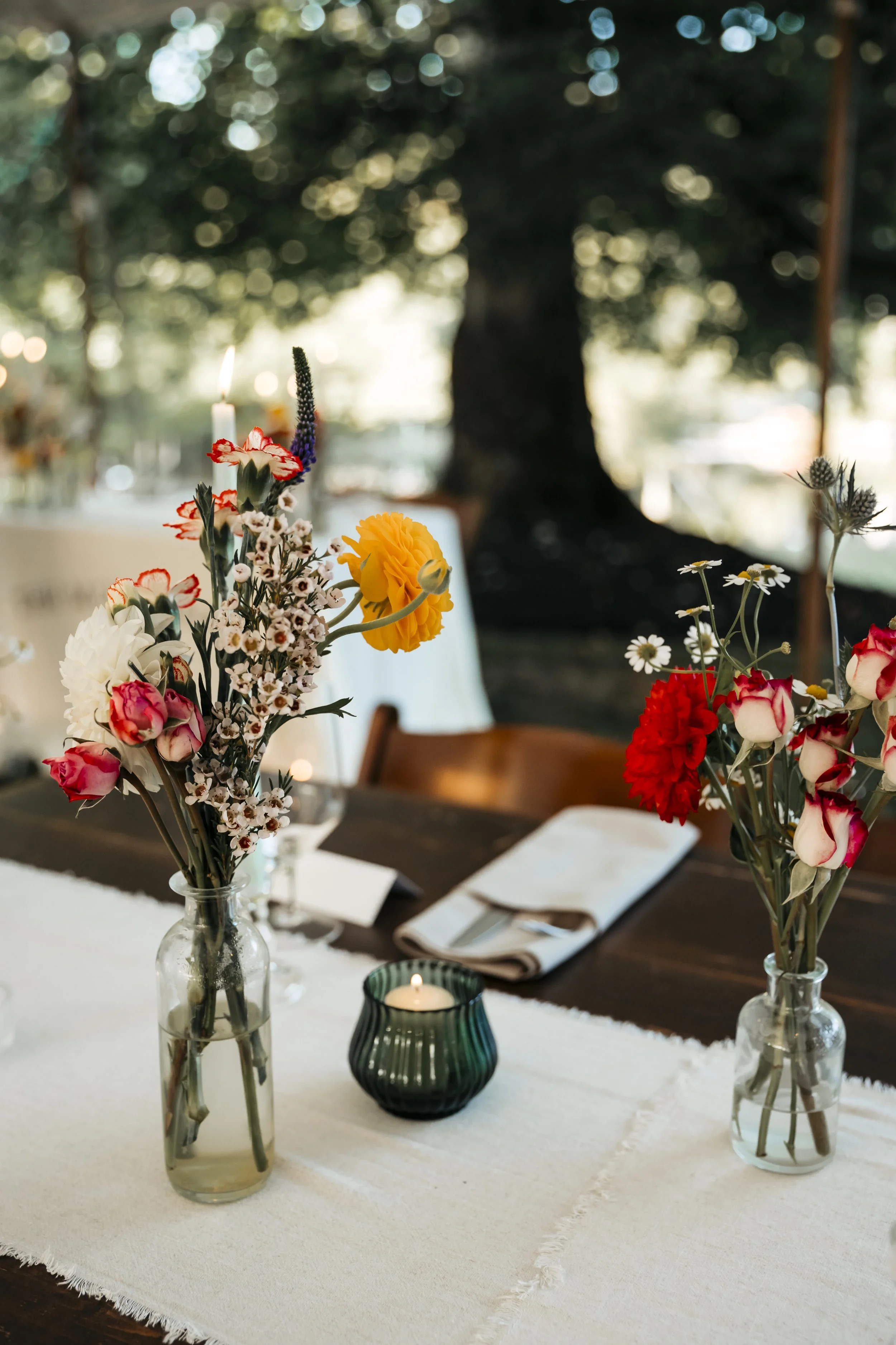 Vase with mixed flowers, candle in green holder, table setting with napkins, outdoors setting with trees and blurred background.