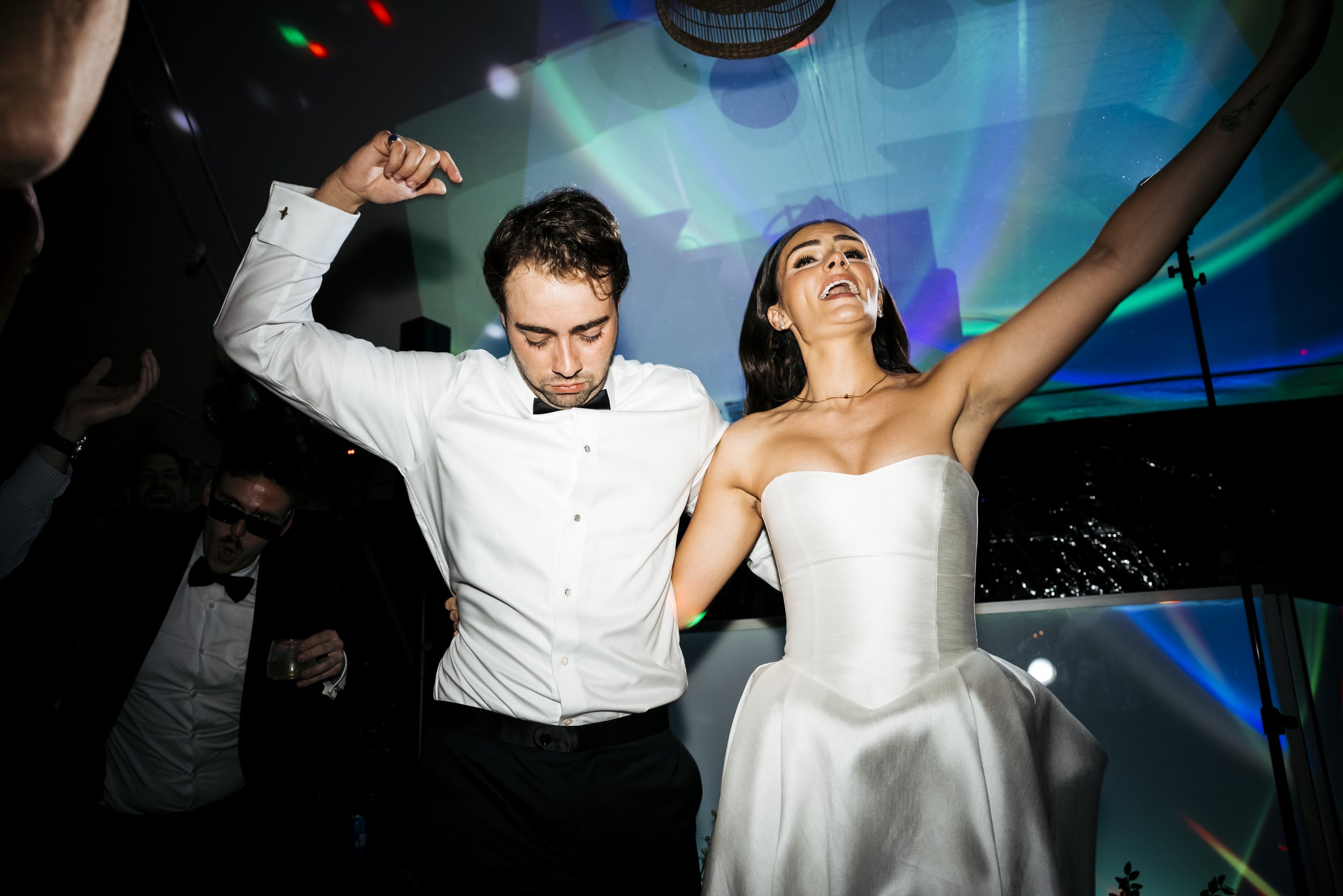 A man and woman dancing at a celebration, with the woman wearing a white strapless wedding dress and smiling with her arms raised, and the man in a white shirt with a black bow tie, dancing with his eyes closed.