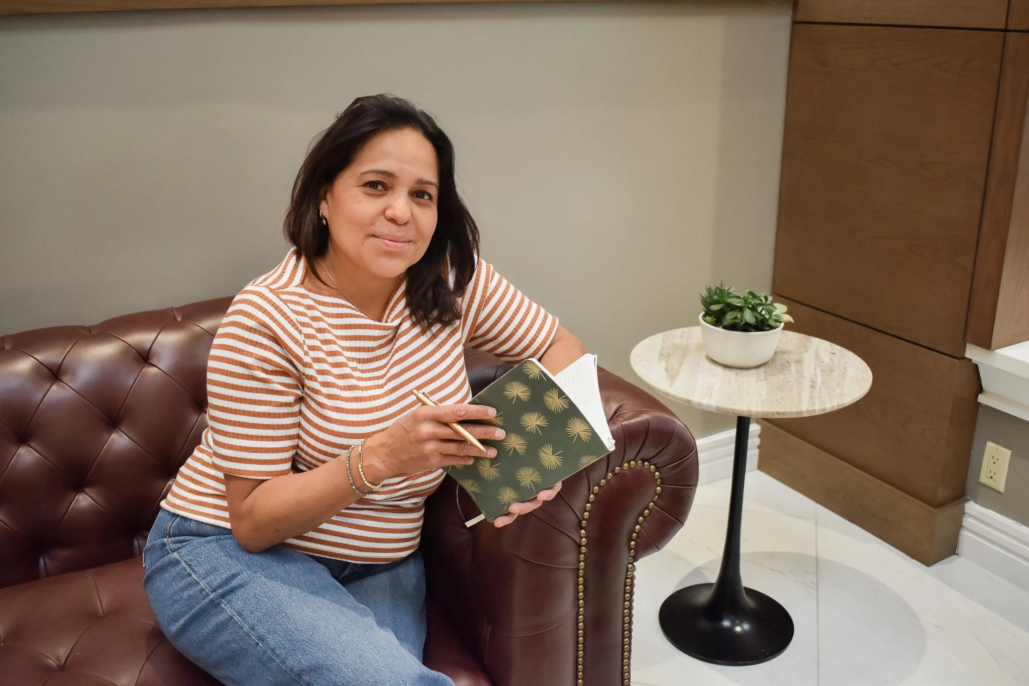 Woman sitting on a brown leather tufted sofa, wearing a striped orange and white top and blue jeans, holding a green notebook with a gold design and a pen, smiling at the camera, with a white marble side table with a potted green plant next to her.