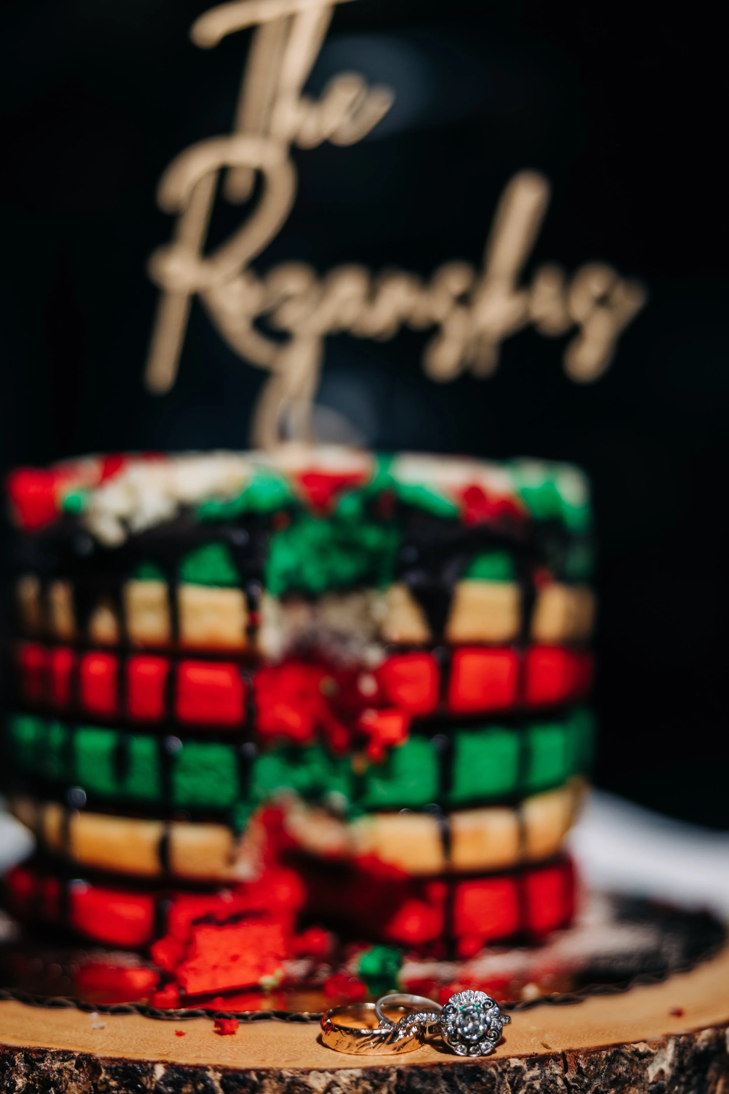 A birthday cake decorated with red, green, and white icing and black drizzle, placed on a wooden surface with a pair of rings in front of it. A sign in the background reads 'The Reardley'.