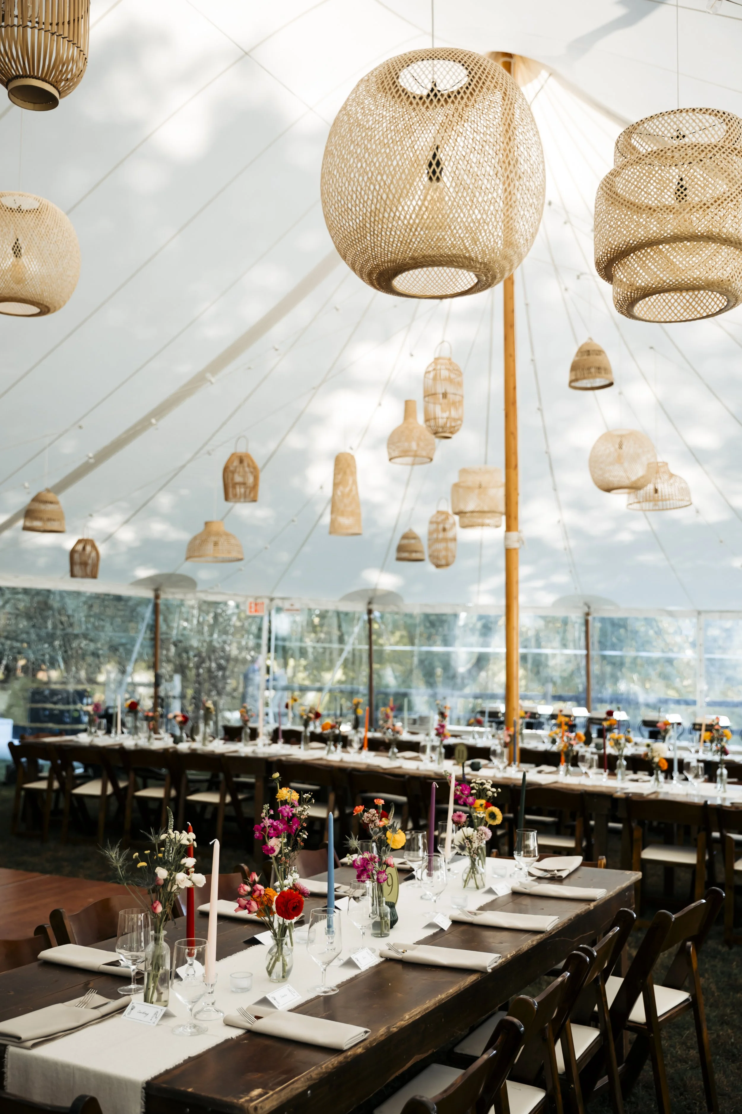 Decorated banquet tent with hanging woven lamps and tables with white tablecloths, floral centerpieces, candles, and glassware.